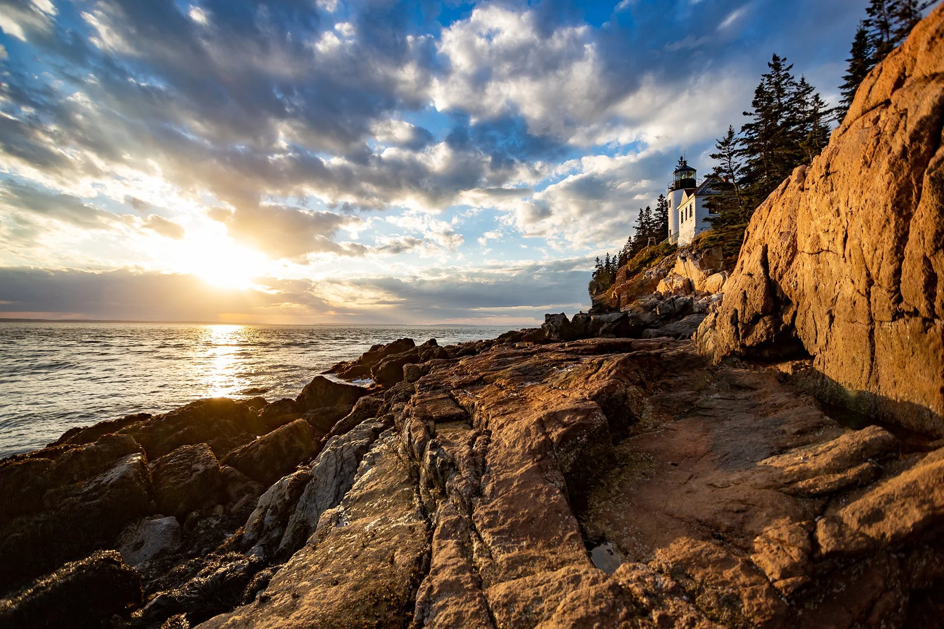 Sunset over the ocean with rocky shoreline and a lighthouse on a hill among trees.