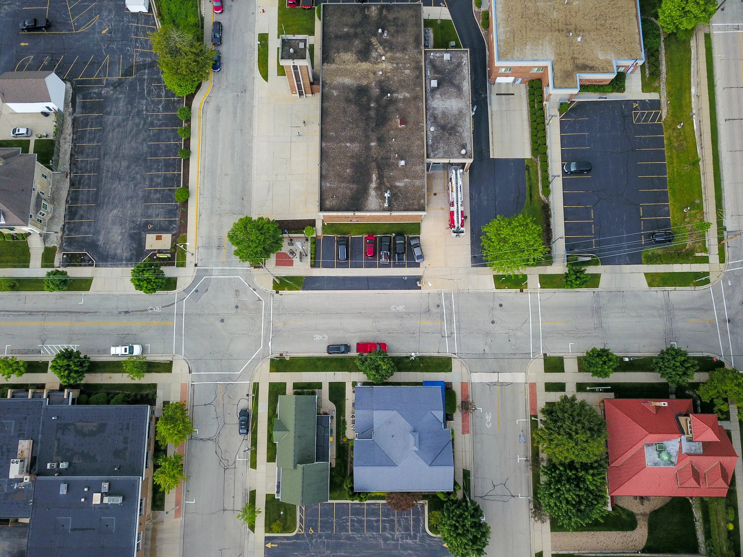 An aerial view of a small town intersection with crosswalks, parking lots, residential houses, trees, and a few vehicles on the streets.