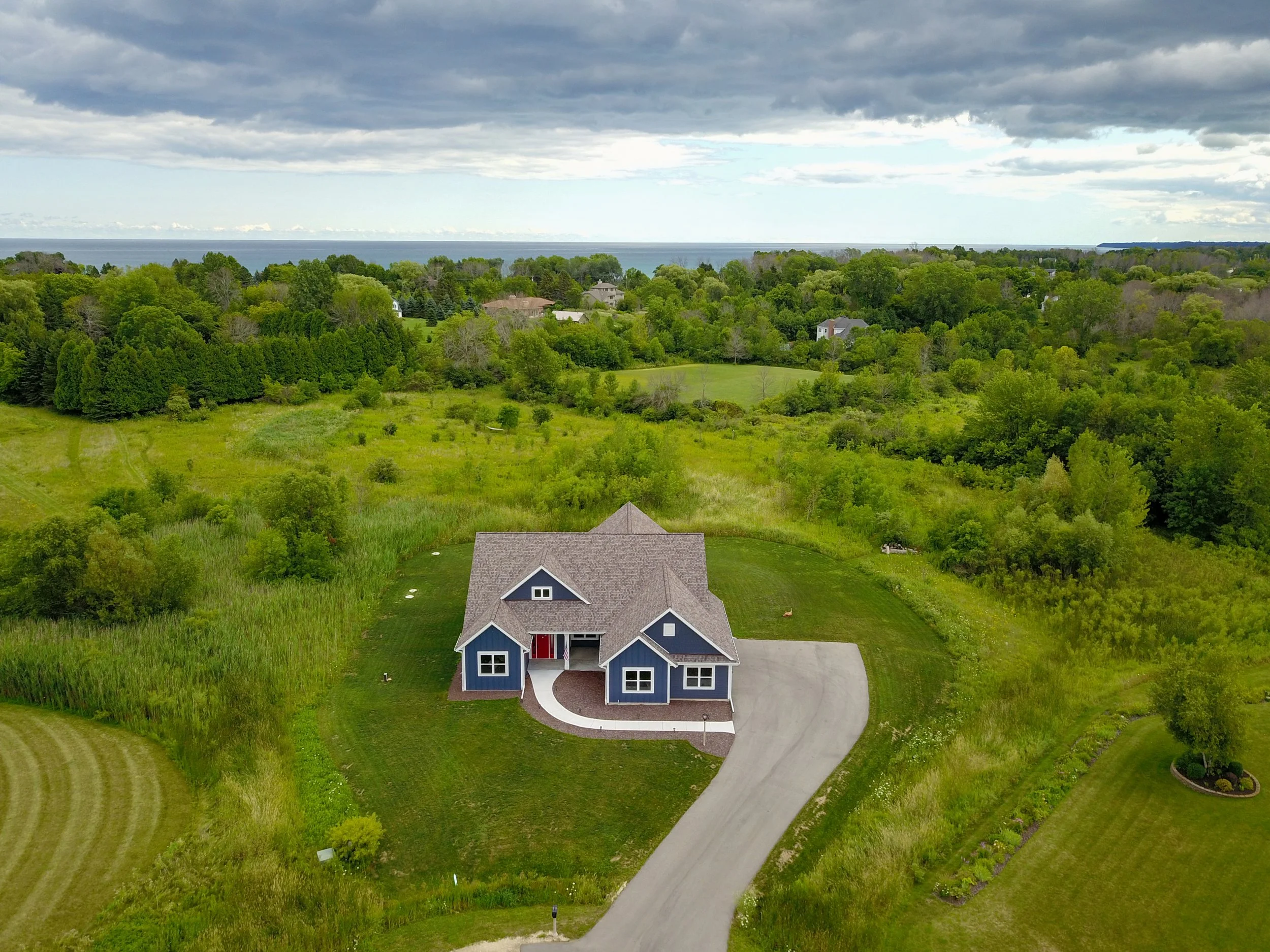 A large blue house with a gray roof, surrounded by lush green lawns and trees, with a curved driveway leading to the front door. In the background, dense green forest extends towards the horizon, with cloudy sky overhead and a body of water visible in the distance.