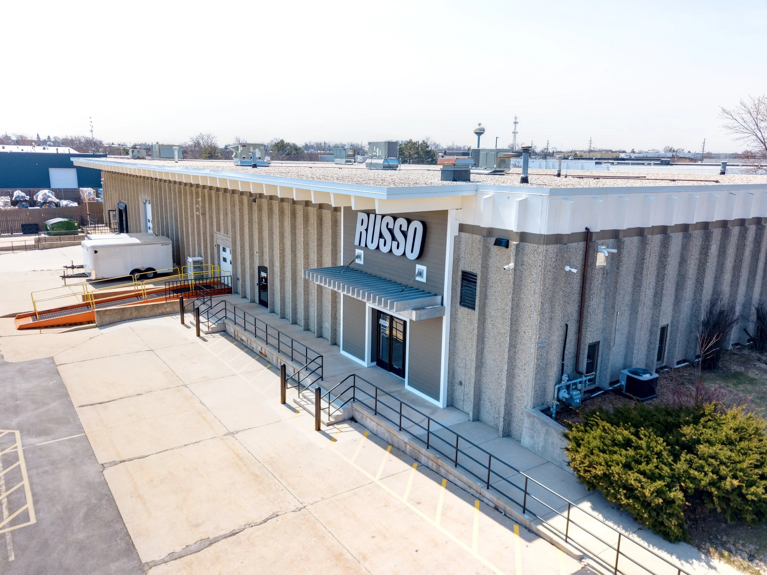 An exterior view of a commercial building with a large sign that reads 'RUSSO', a concrete parking lot with marked parking spaces, a trailer, and a ramp with yellow rails, under a clear sky.