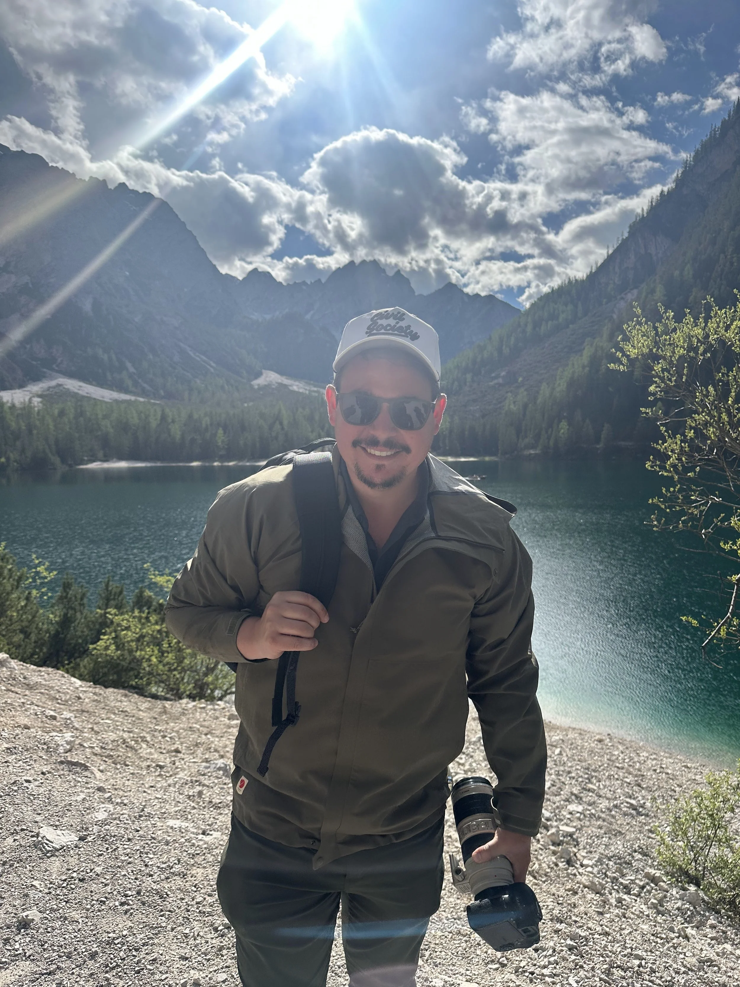Wesley Wahlberg smiling, wearing sunglasses, a hat, and a jacket, holding a camera, standing near a lake in Italy with mountains and clouds in the background under a bright sun.
