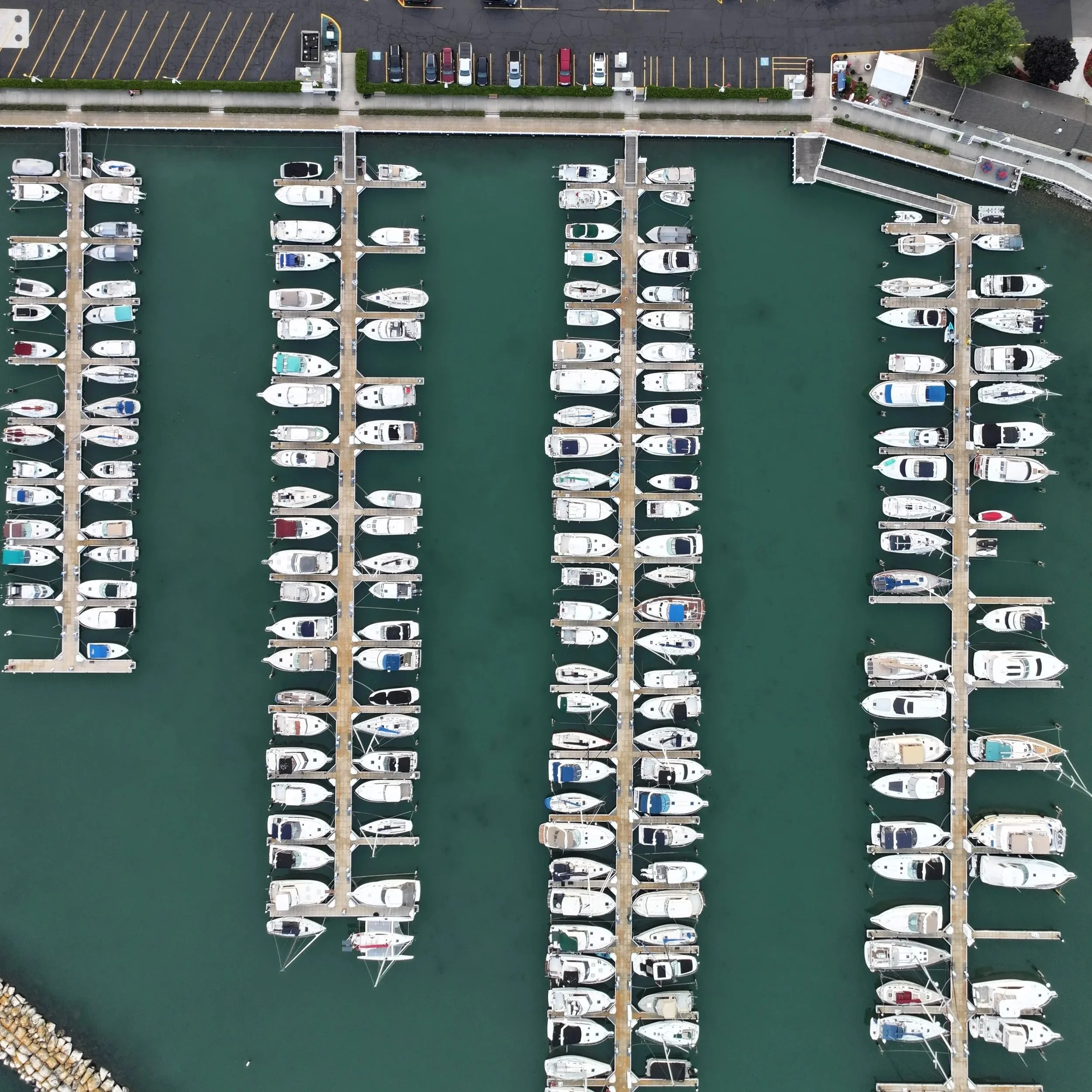 Aerial view of a marina with multiple rows of docked boats and yachts in the water, with parking lot and some trees visible at the top.