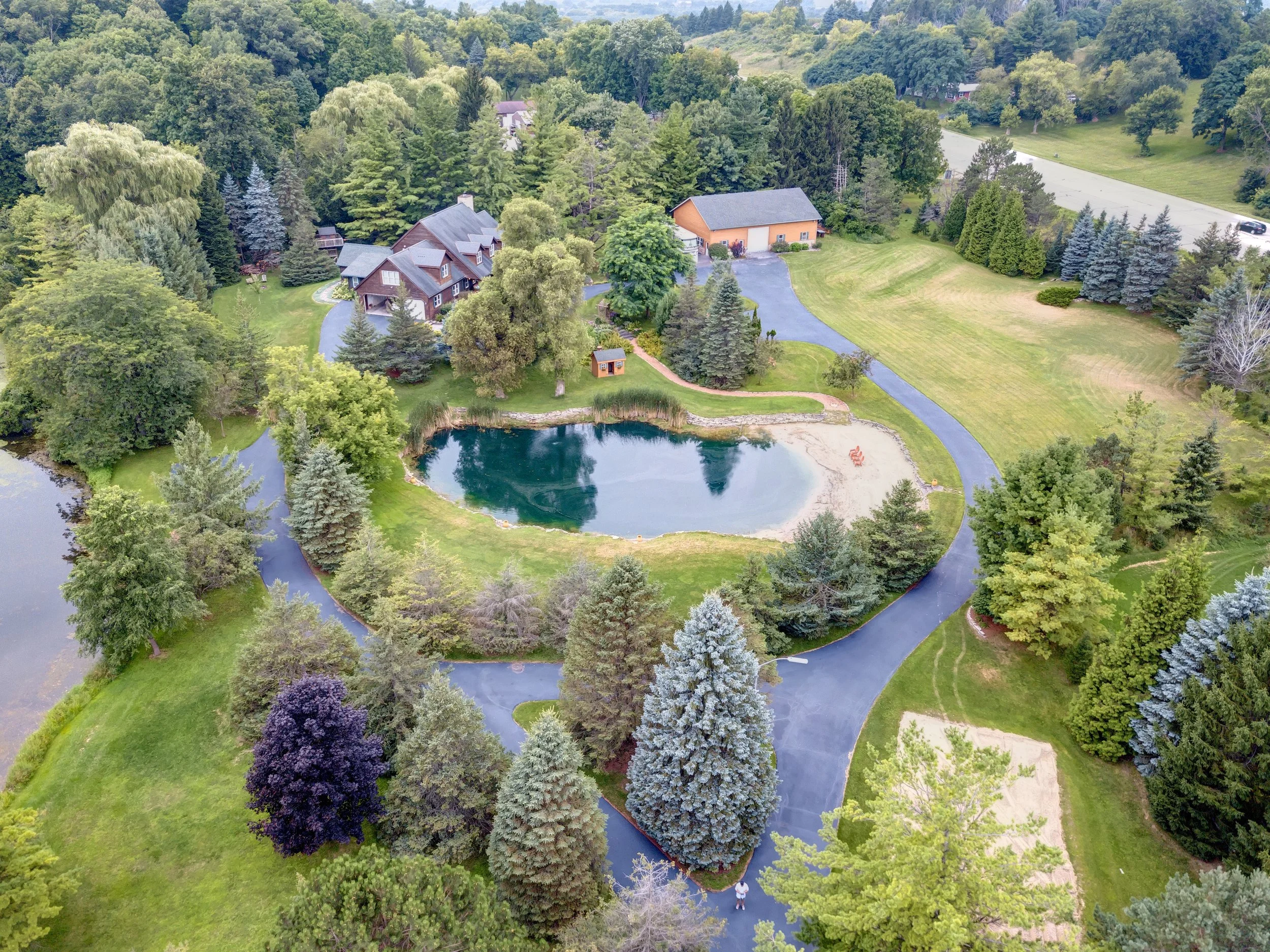 Aerial view of a property with a pond, surrounded by trees, a house, garage, and a driveway in a lush green landscape.