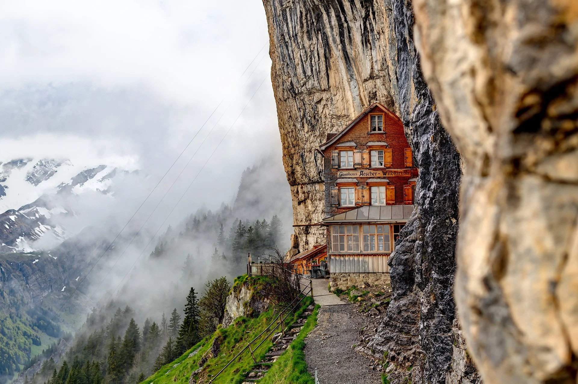 A house built into the side of a rocky mountain cliff with a forested valley and snow-capped mountains in the background, under cloudy sky.