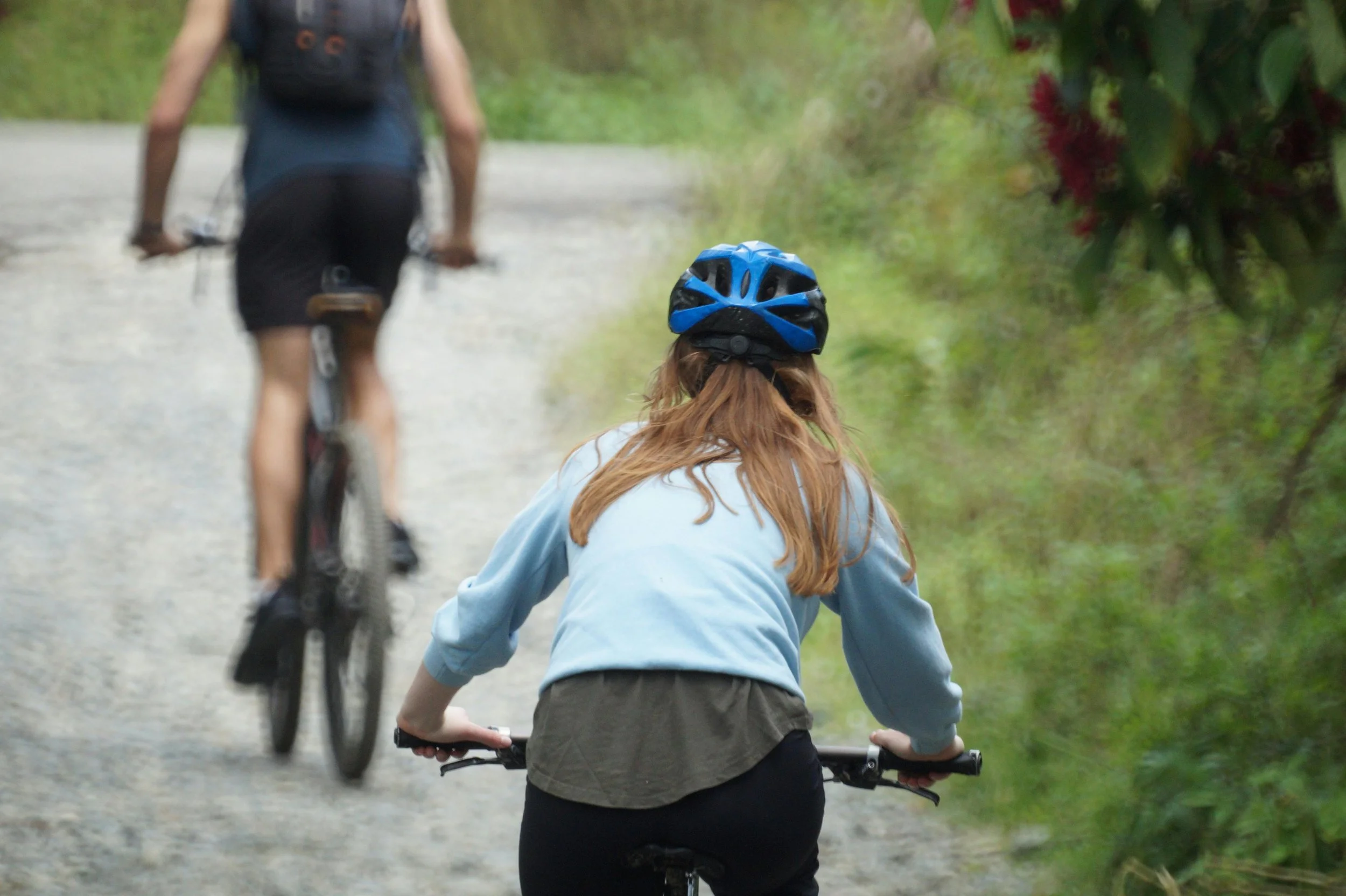A person with long red hair wearing a blue helmet riding a bicycle on a dirt trail, with another cyclist and greenery in the background.
