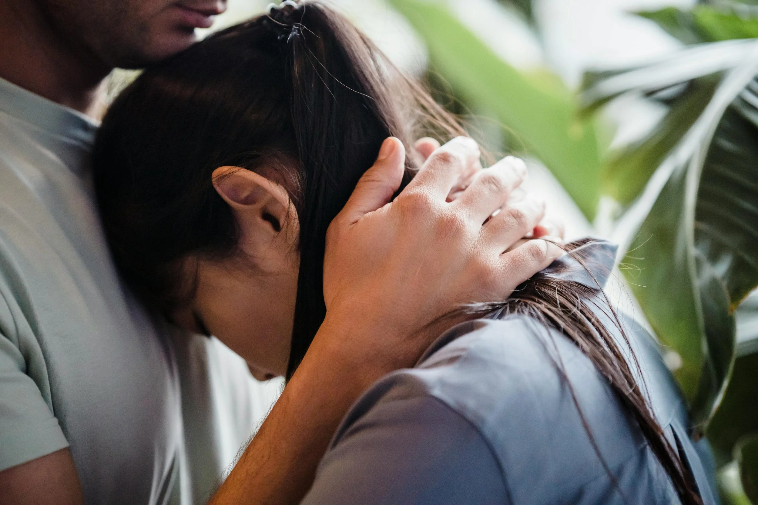 A person with dark skin gently comforting or consoling a person with lighter skin and long dark hair by holding their head and resting their forehead against theirs. They are indoors with blurred green plants in the background.