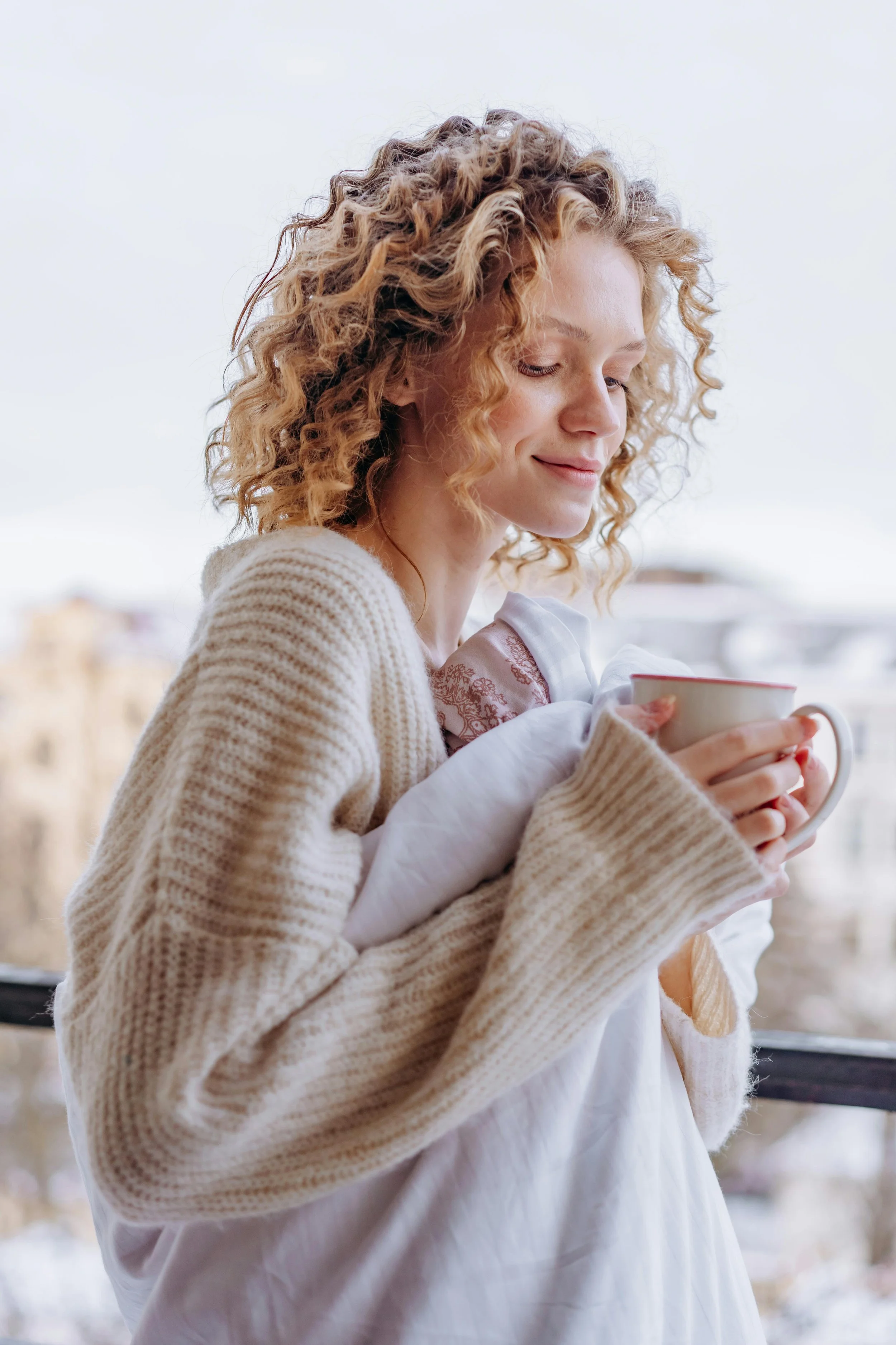 A woman with curly red hair is holding a white mug and gazing down with a gentle smile.