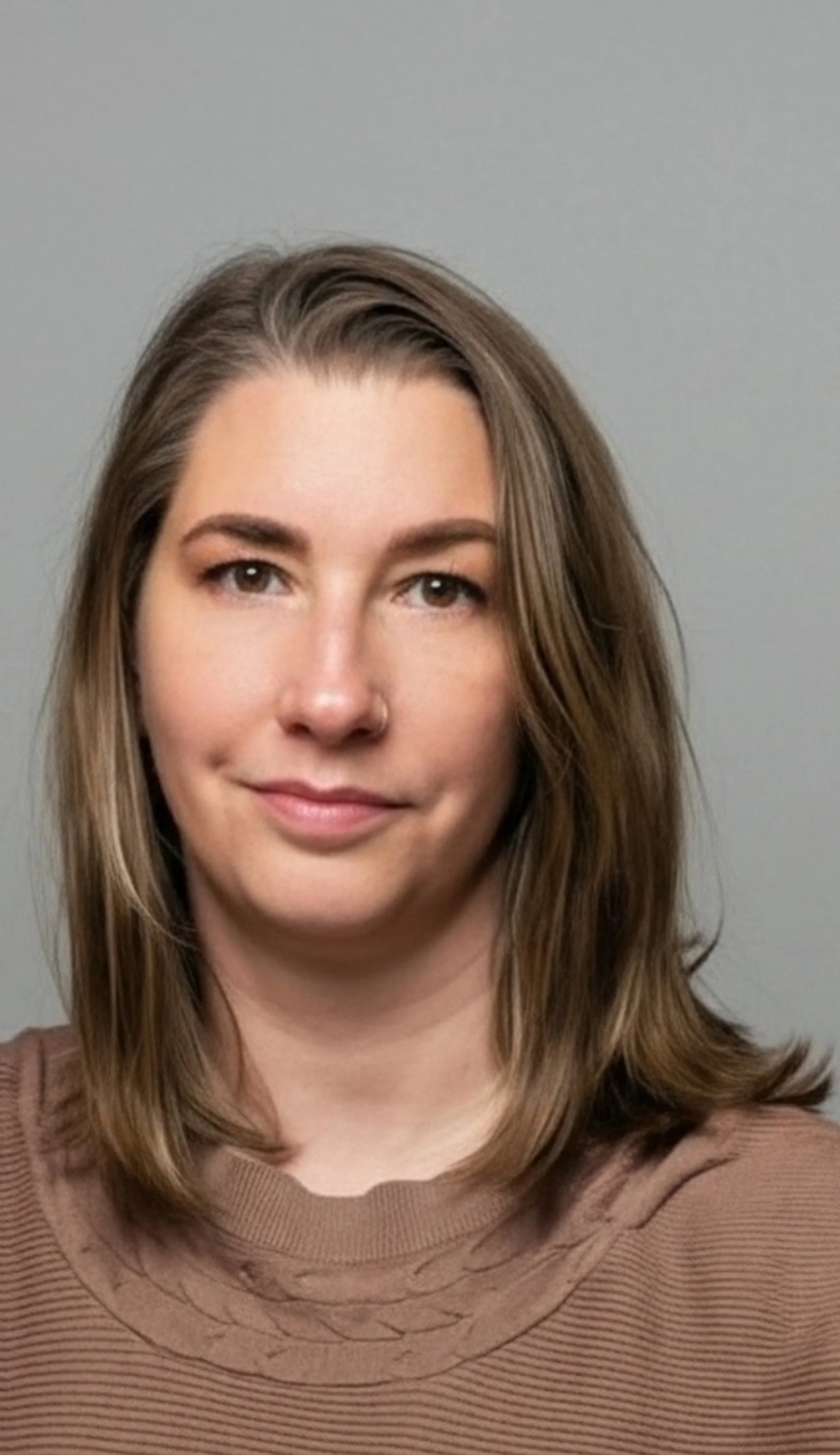 A woman with shoulder-length brown hair, wearing a brown top, smiling slightly, against a plain gray background.