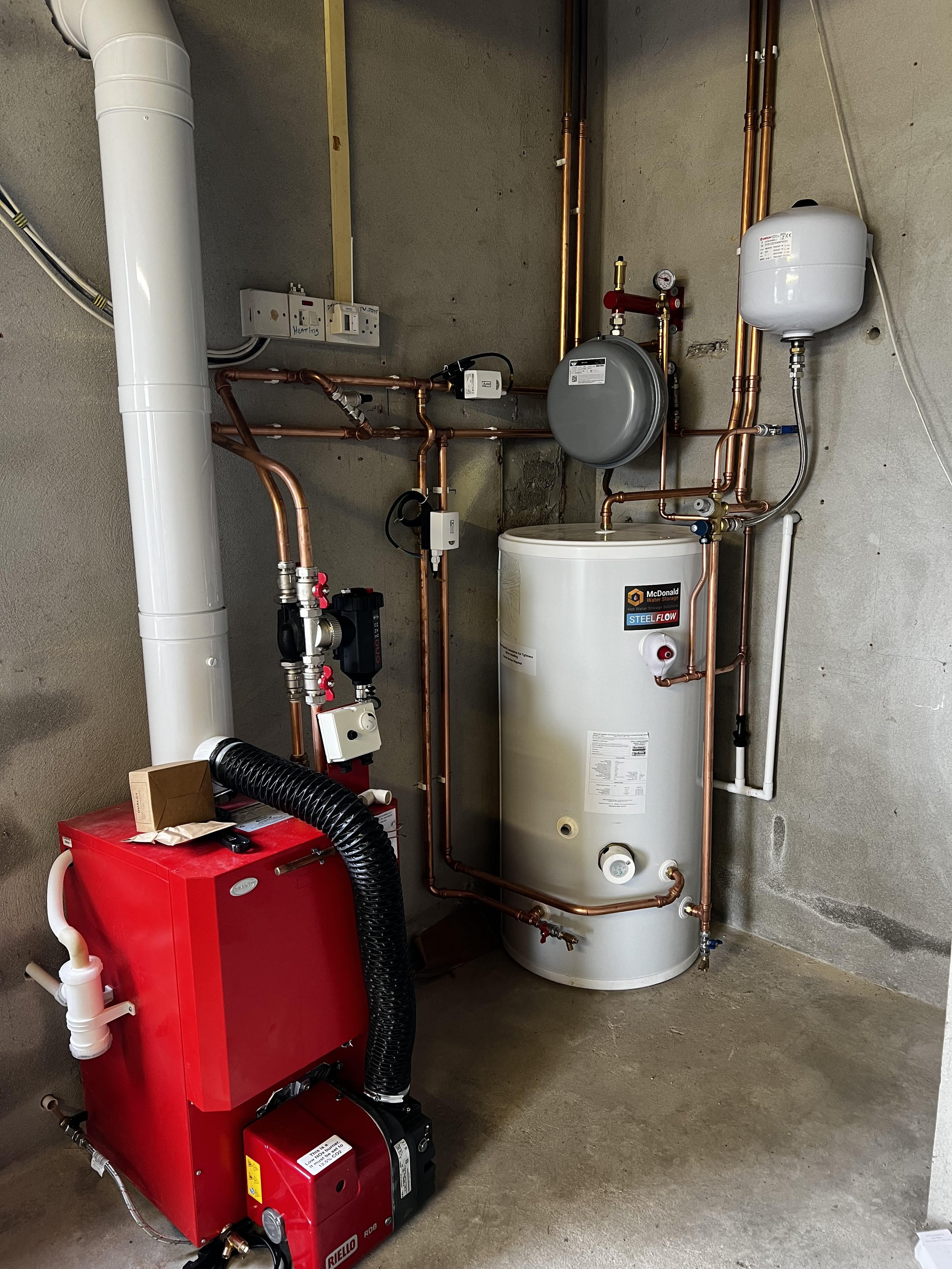 A utility room with a large water heater, copper pipes, a red boiler, and various plumbing and electrical components against a concrete wall.