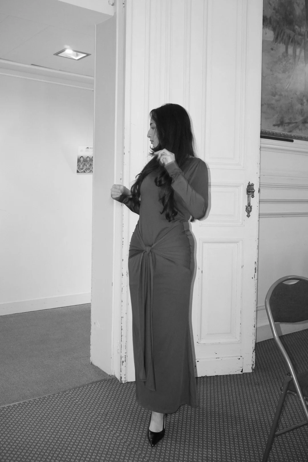 Femme en robe longue, regardant vers la gauche, comme posant, à l'intérieur d'une pièce avec porte en bois et mur blanc.