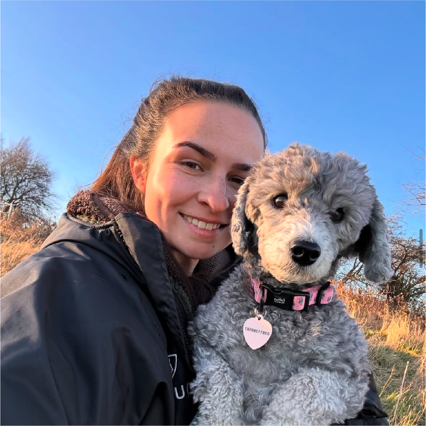 A woman smiling outdoors with a gray curly-haired puppy, both facing the camera, during sunset with trees and grass in the background.
