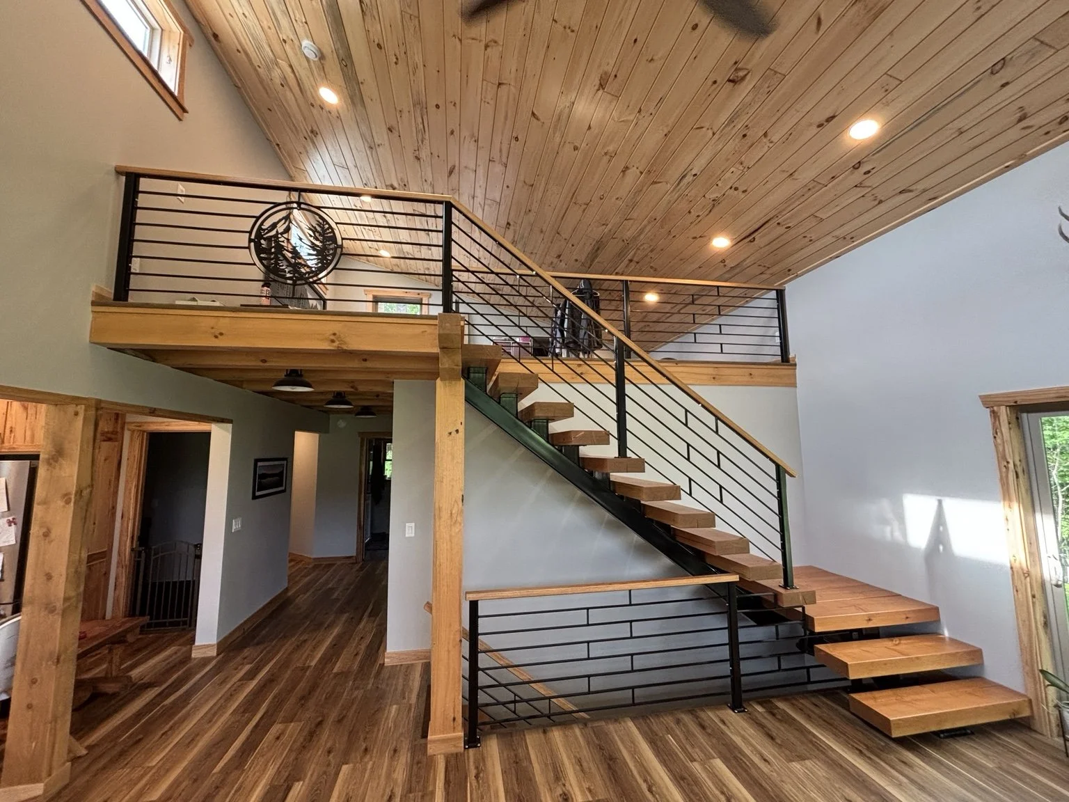 A modern two-story house interior featuring a staircase with wooden steps and black metal railings, a wooden ceiling, and hardwood floors, with an upper balcony area visible.