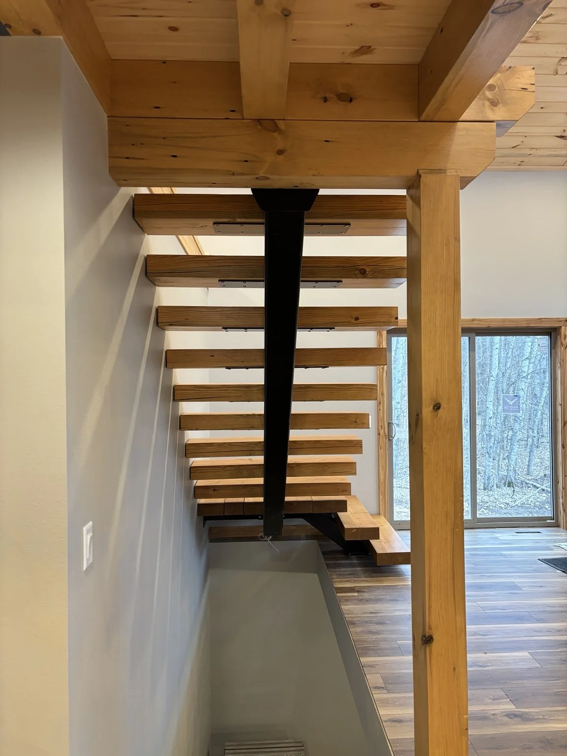 Interior view of a modern wooden staircase with open risers, black central support beam, leading to an upper level in a home with wooden flooring and large window showing trees outside.