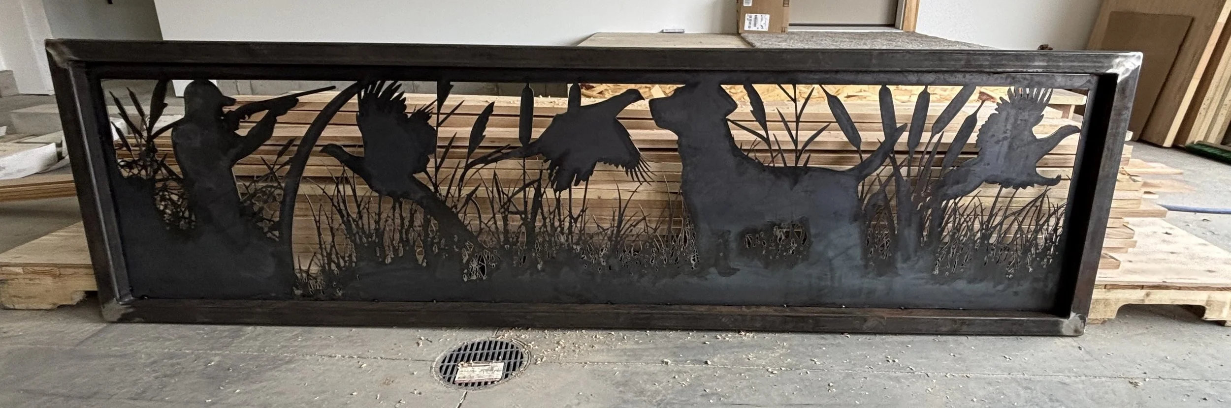 A rectangular metal wall art piece depicting a nature scene with birds and a deer among tall grasses, set against a wooden background.