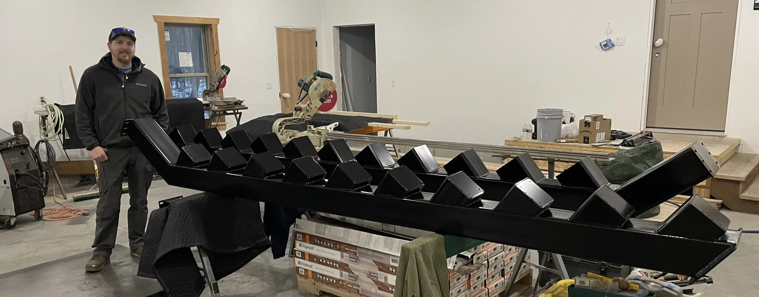 A man standing in a workshop next to a large black metal structure of an inclined ladder with multiple steps.