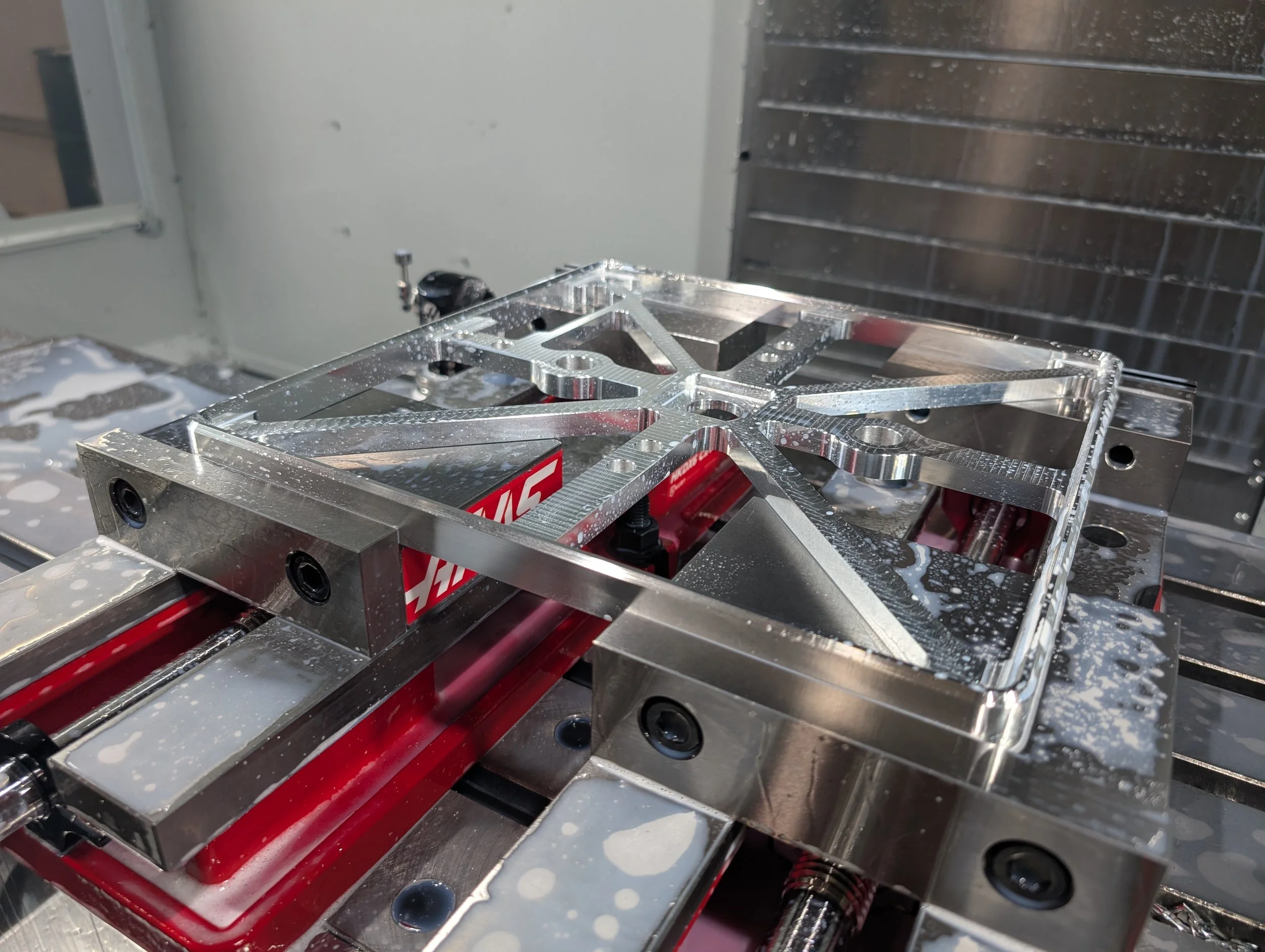 Close-up of a metal fixture and red machinery part on a machine bed, with water splashes and foam, in a manufacturing or workshop setting.