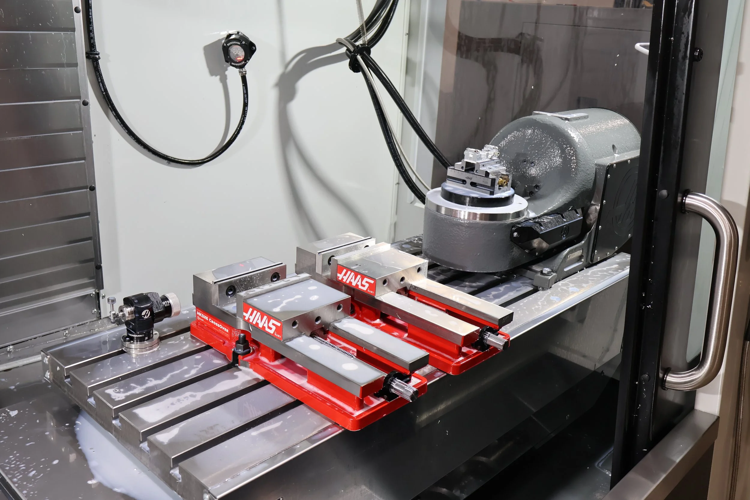 A robotic manufacturing or testing machine with metal components, hydraulic or pneumatic equipment, and red fixtures labeled HAAS in a clean industrial setting.