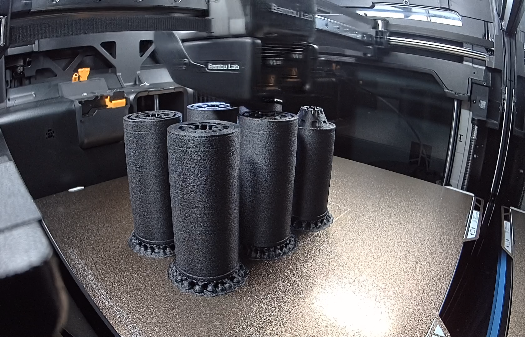 Four black cylindrical 3D printer filament rollers on a textured surface inside a 3D printer enclosure.