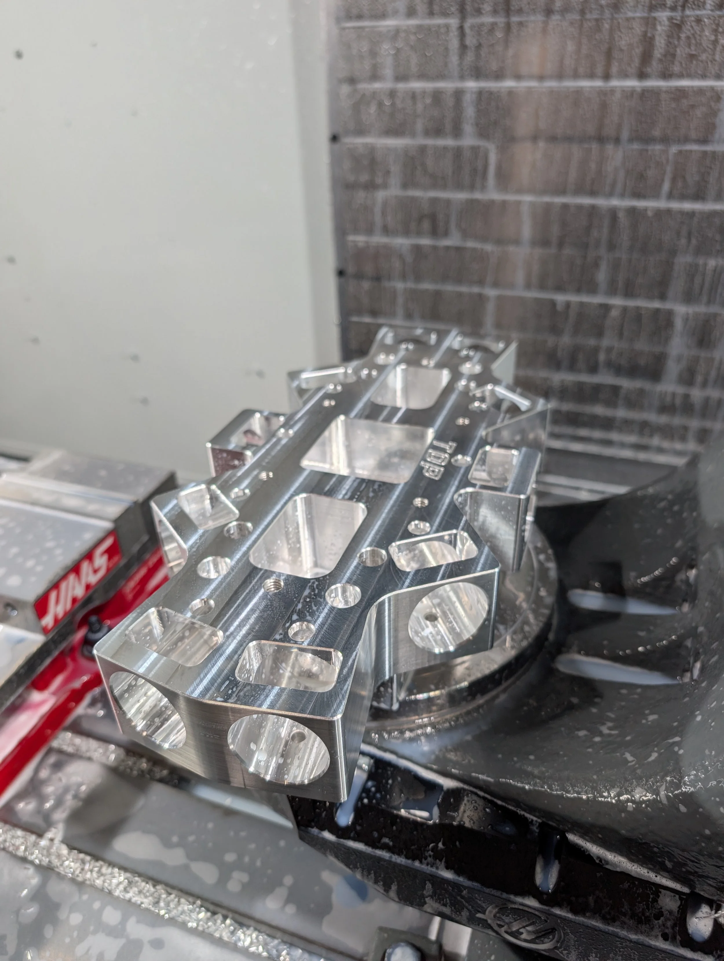 Close-up of a machined metal component on a CNC machine, with a brick wall in the background.