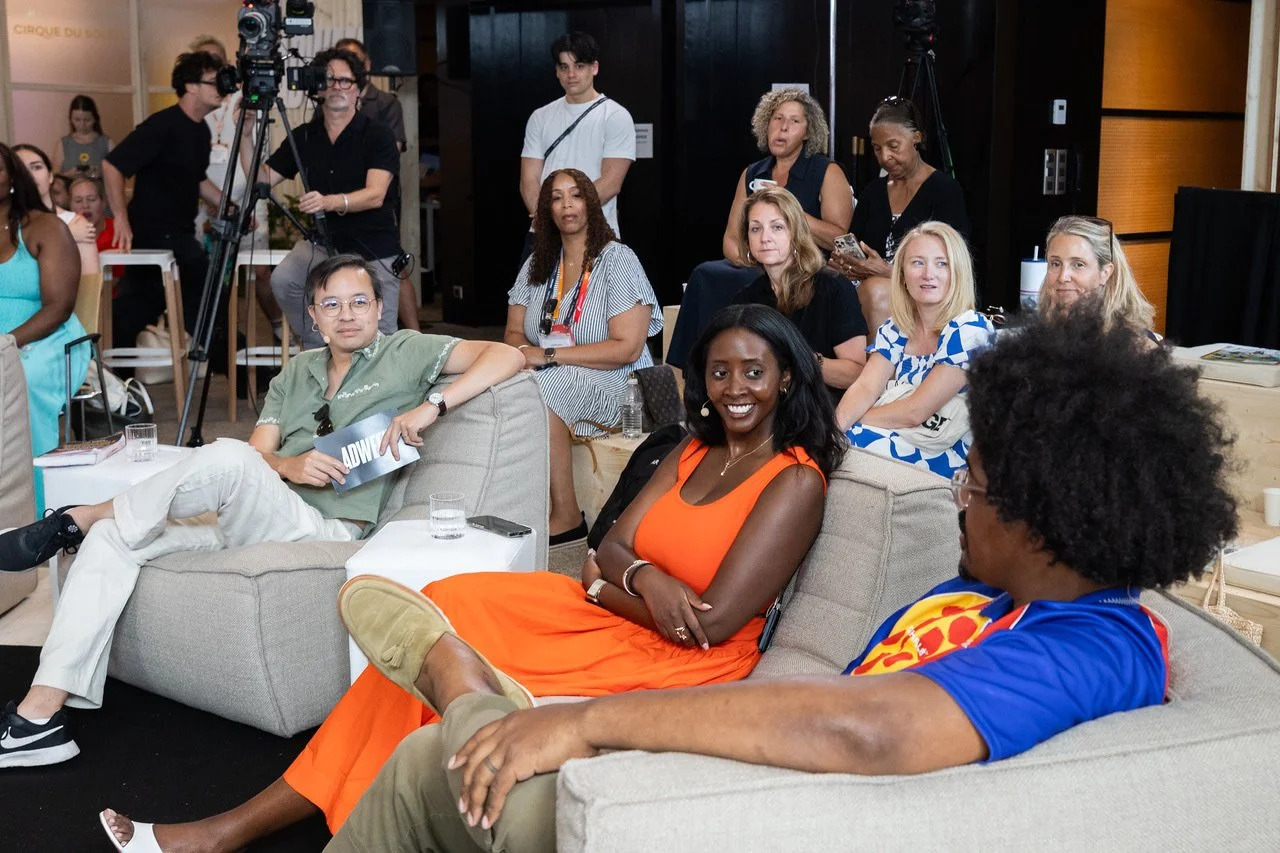 Group of diverse people sitting on couches and chairs at an indoor conference or event, some smiling and engaging, with cameras and equipment in the background.