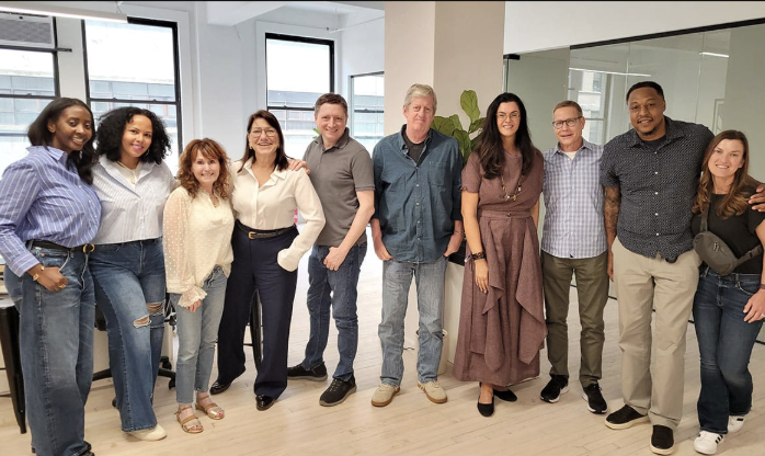 Group of eleven diverse people standing together in a modern office, smiling at the camera.