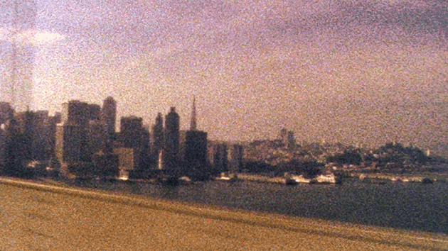 City skyline with skyscrapers near water, viewed from a moving vehicle.