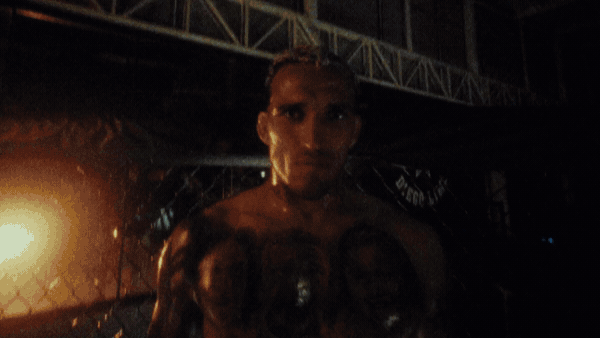 A woman with a serious expression, covered in tattoos, standing in a dark industrial or stage-like setting with metal structures and orange lighting in the background.