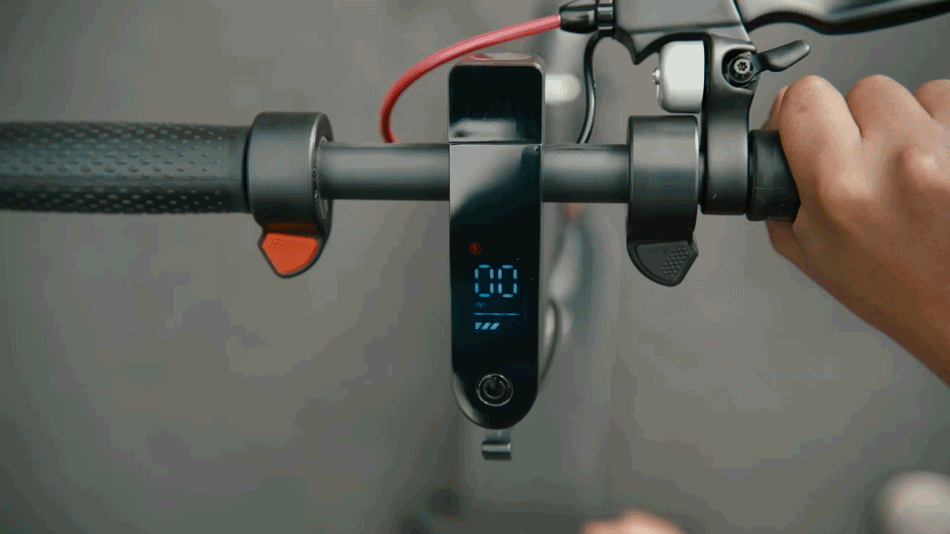 Close-up of a digital display on an electric scooter's handlebar, showing the speed and battery level, while a hand grips the right handle.
