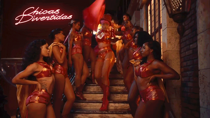 Group of women in shiny red costumes standing on and around staircase in a dimly lit room, with a neon sign reading 'Chicas Sinverguenzas' in the background.