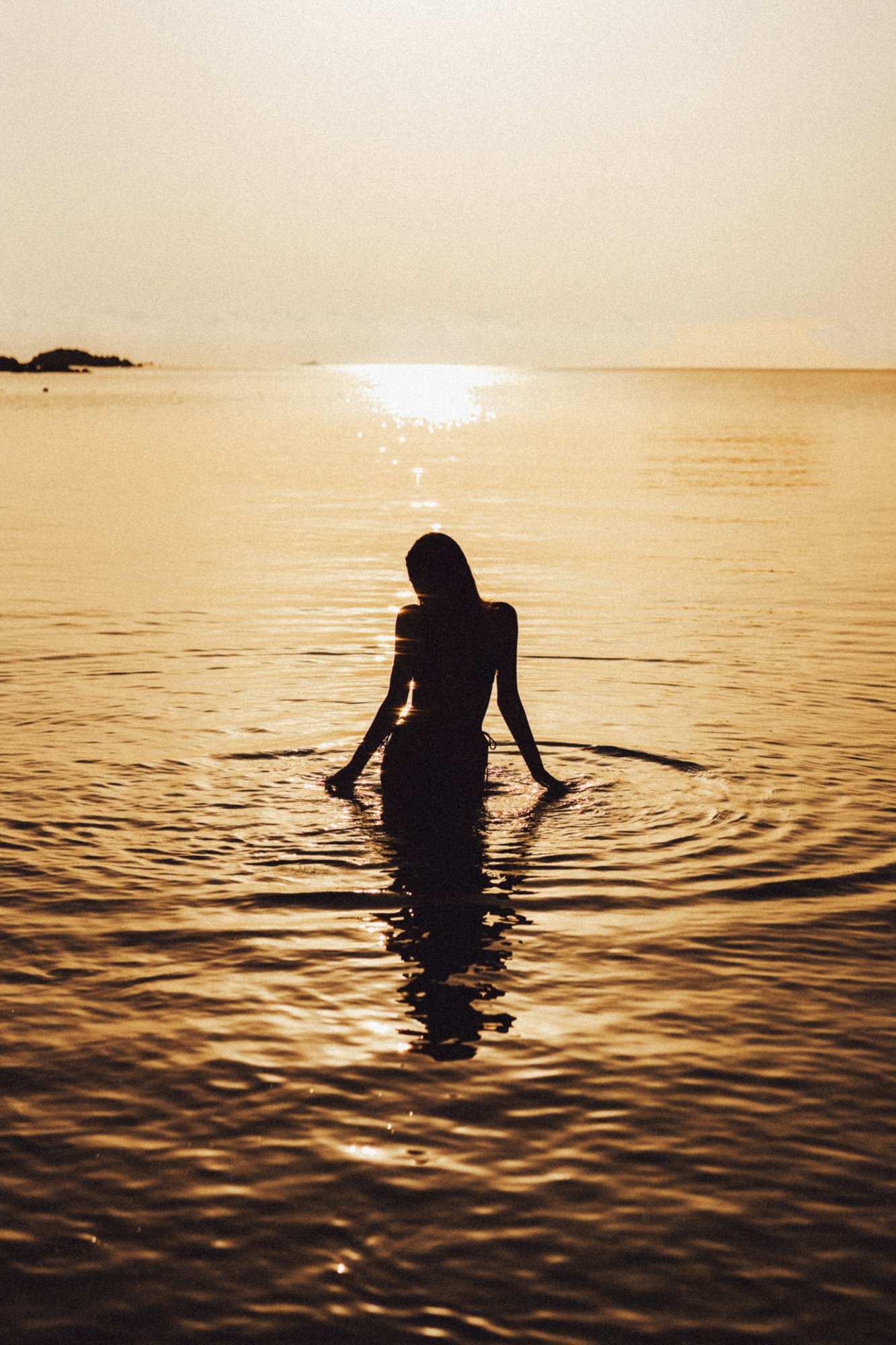 Una donna seduta nell'acqua durante un tramonto, con il sole che si riflette sul mare.