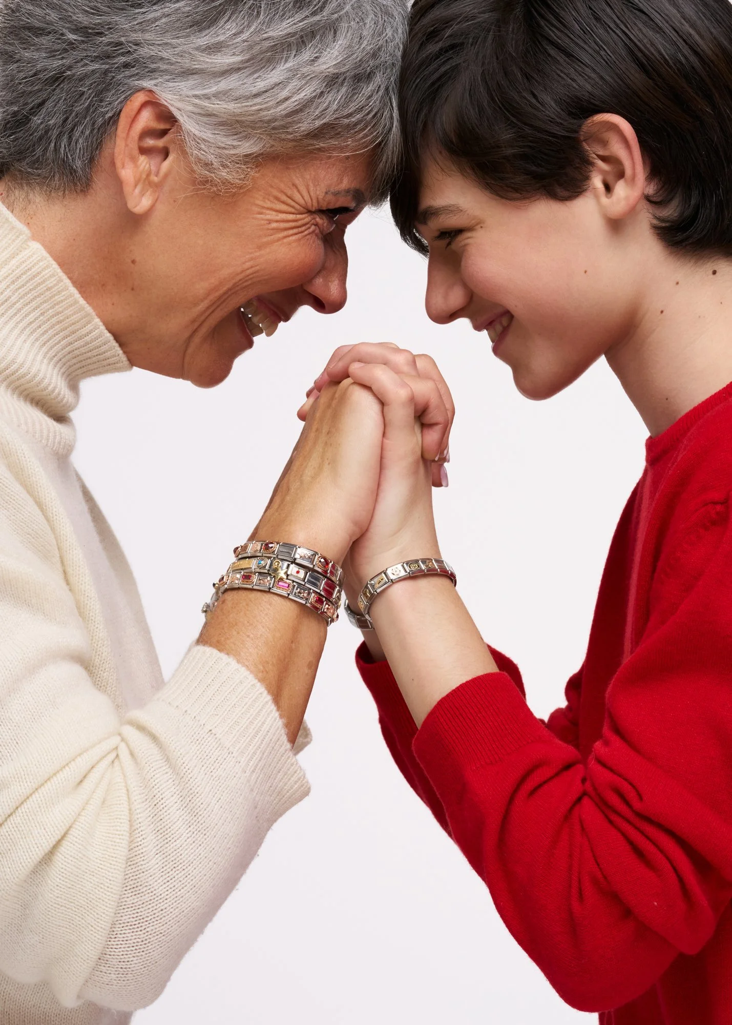 Due donne, una anziana con capelli grigi e l'altra giovane con capelli castani, si fronteggiano sorridenti e si tengono per mano, con le testa chine e un sorriso complice.