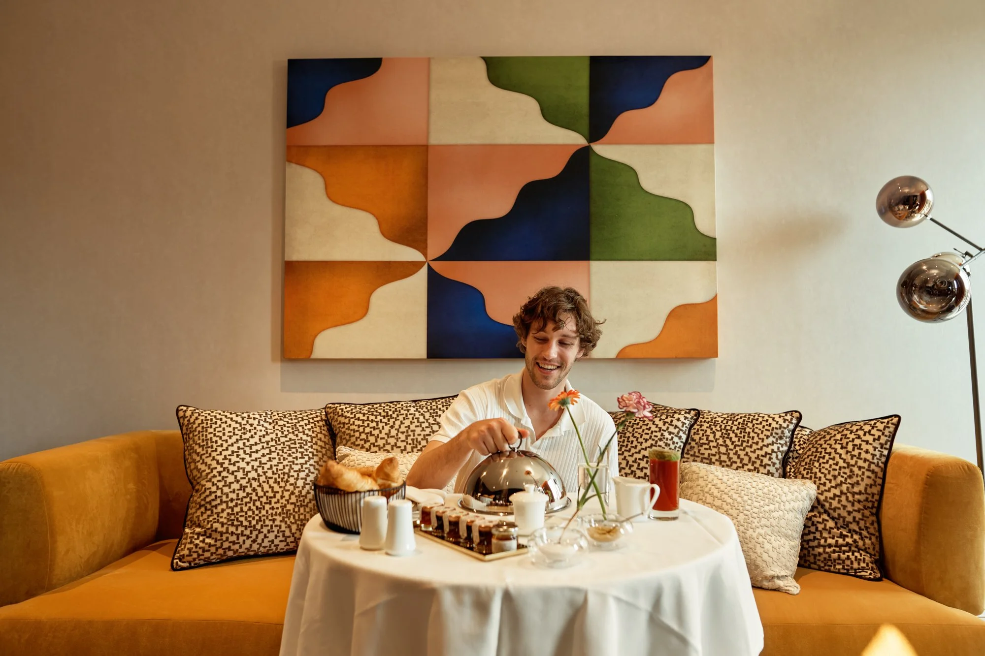 Un ragazzo con capelli ricci, sorridente, che si sta godendo una colazione con tè, dolci e fiori in un ambiente accogliente con dipinto astratto alle spalle.