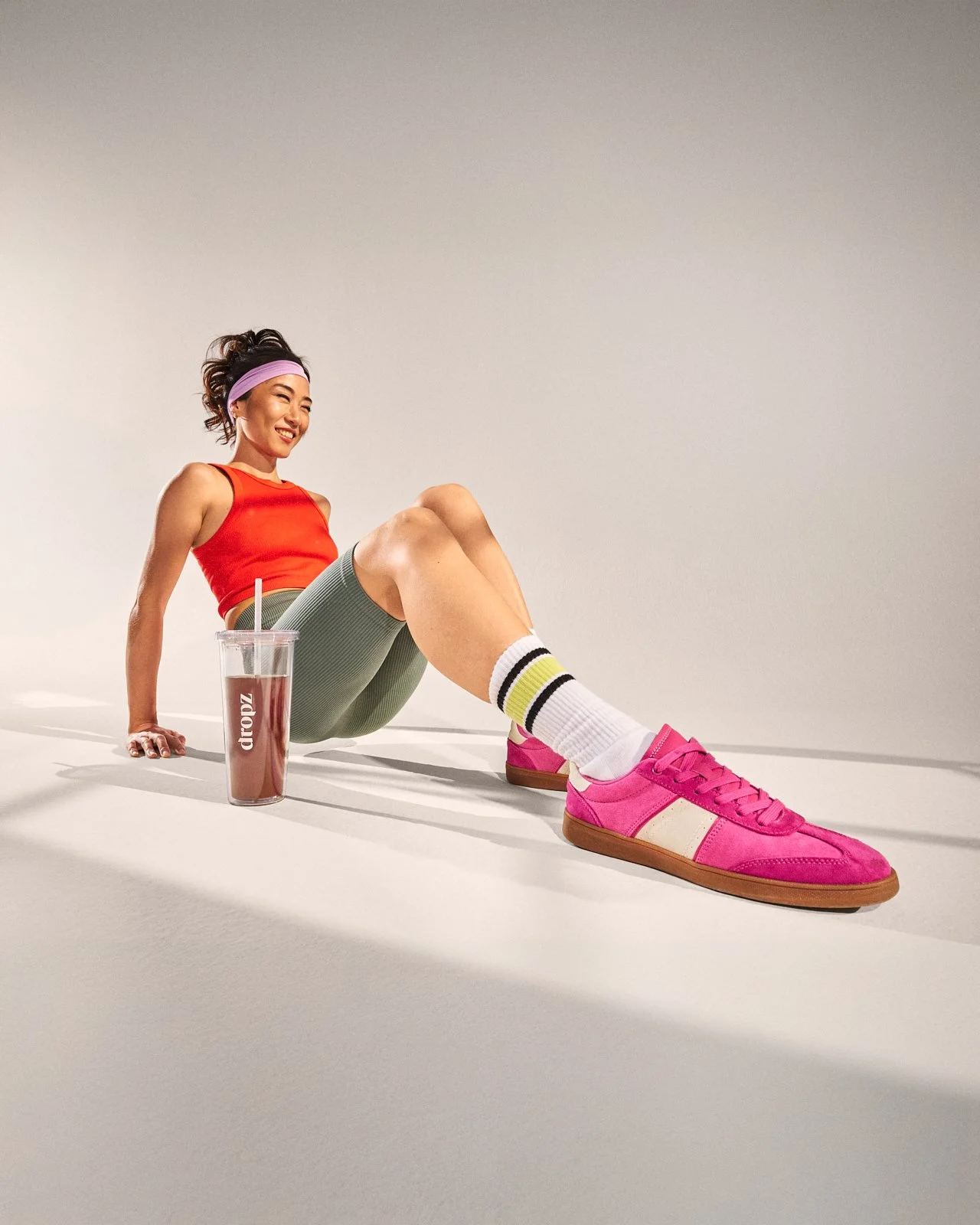 Una donna sorridente in abbigliamento sportivo seduta a terra con una grande borraccia rosa, indossa una maglietta rossa, collant grigi, calzini bianchi con dettagli nero e giallo, e scarpe da ginnastica rosa.