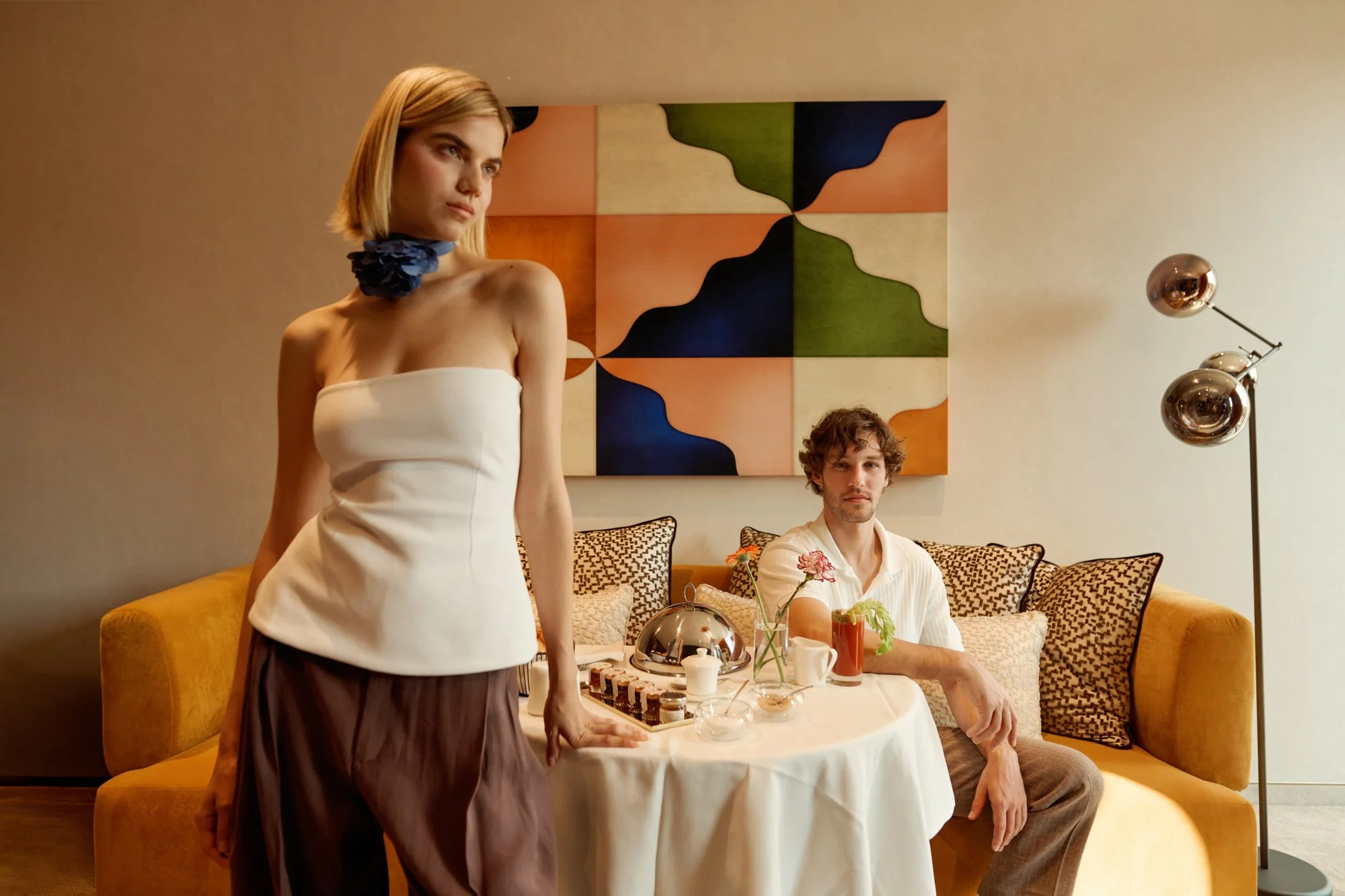 Una donna con capelli biondi e vestito bianco si trova davanti a un uomo con capelli ricci e camicia bianca seduto a un tavolo apparecchiato con vari alimenti, in un soggiorno con parete beige, decorazioni geometriche e luci a soffitto.