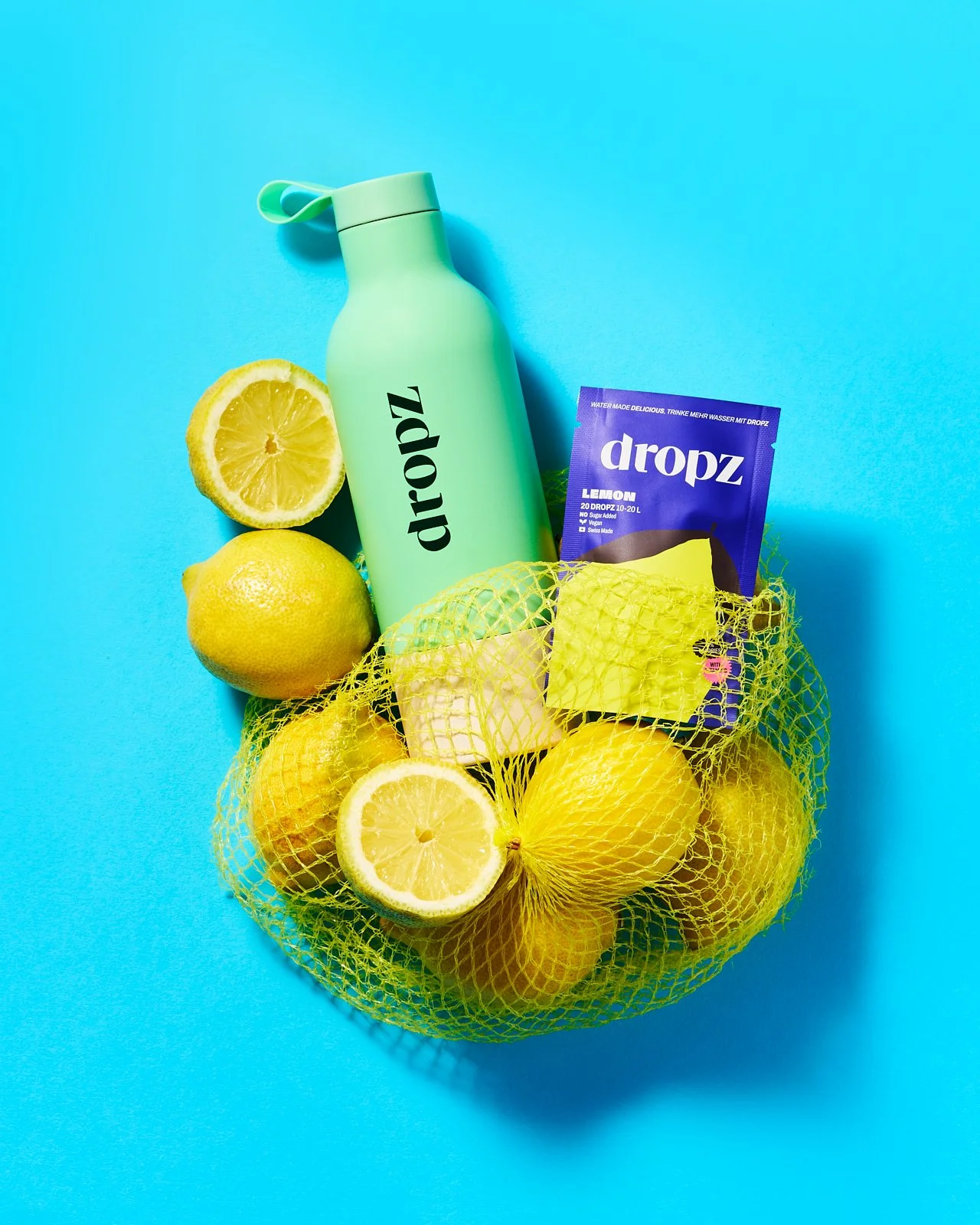 Sacchetto giallo di limoni, limoni freschi e spremuti, bottiglia verde di dropz limone, pacchetto di zuccheri arancione su sfondo blu.