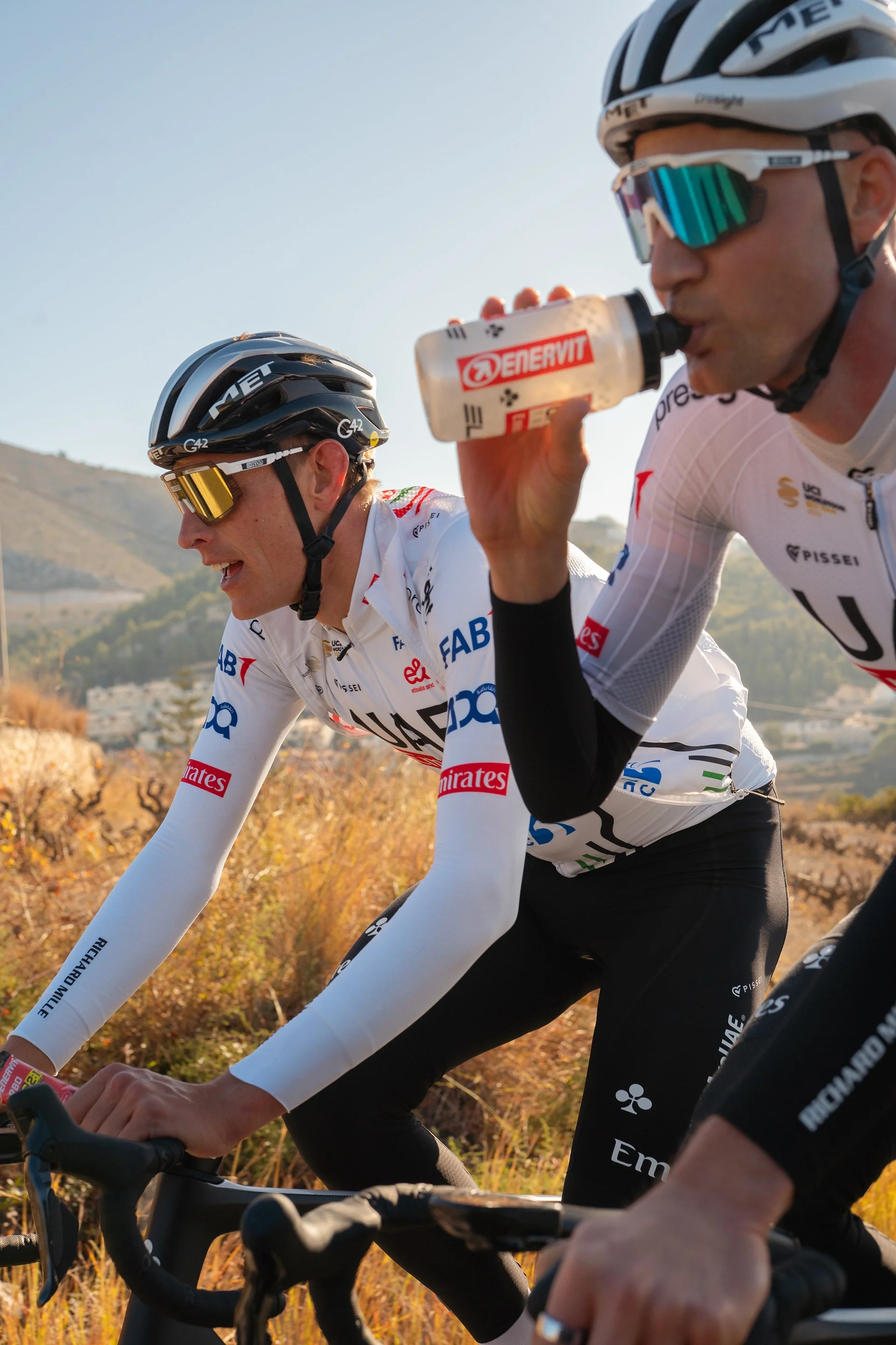 Due ciclisti professionisti in tenuta da corsa, con caschi e occhiali, pedalano lungo un percorso di campagna soleggiato. Uno dei ciclisti beve da una borraccia con il logo Enervit.