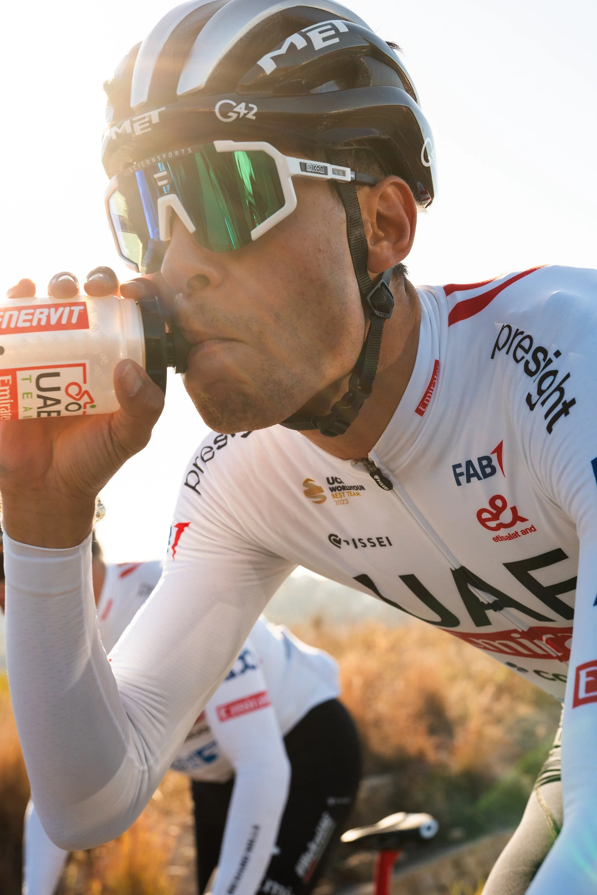 Un ciclista professionista si sta bevendo una bevanda durante una competizione ciclistica su un percorso all'aperto, con un paesaggio naturale sullo sfondo.