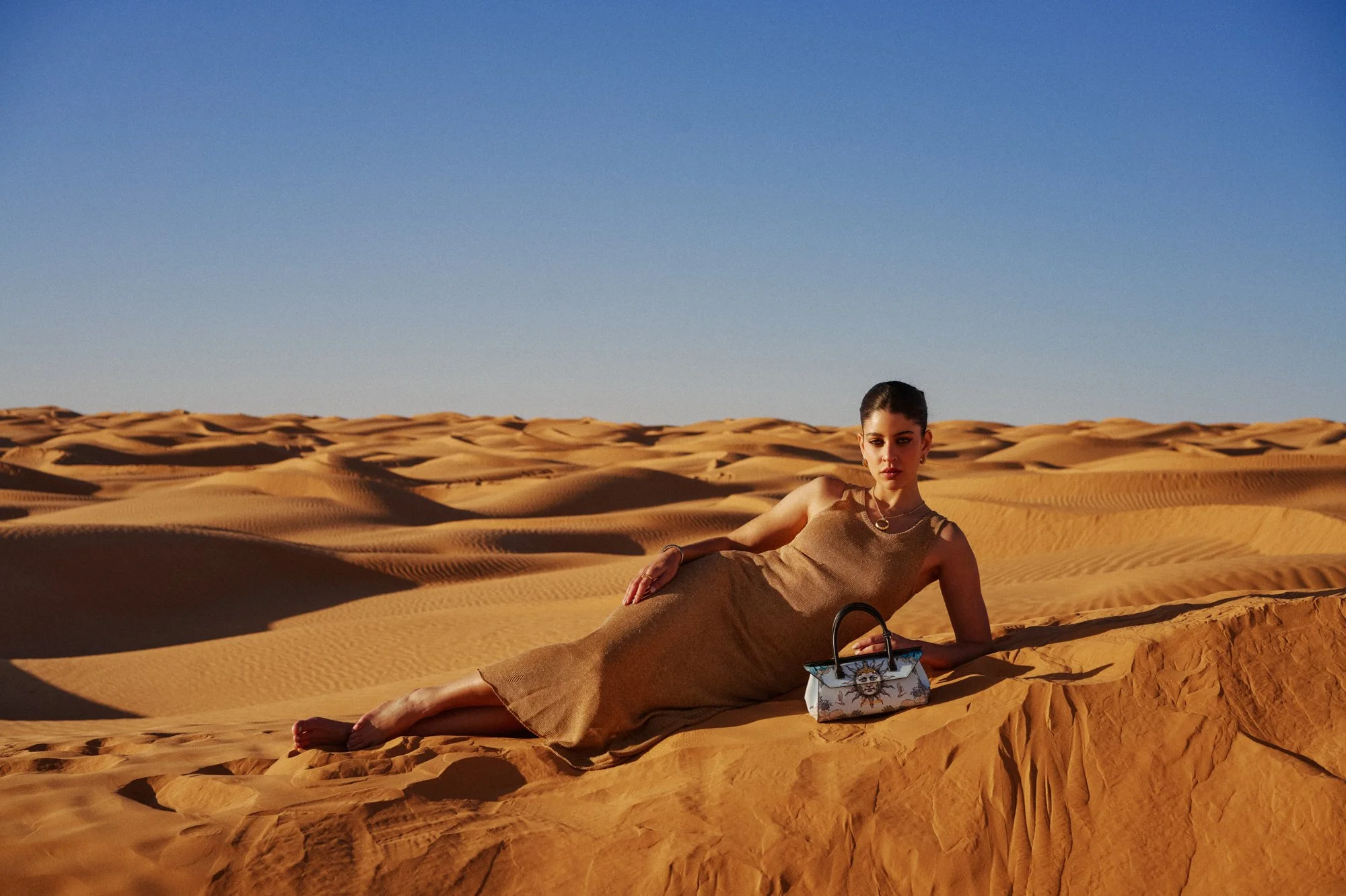 Una donna distesa sui sand dune del deserto con un vestito beige, posando con un accessorio colorato e una borsa decorata. Sotto un cielo azzurro chiaro.