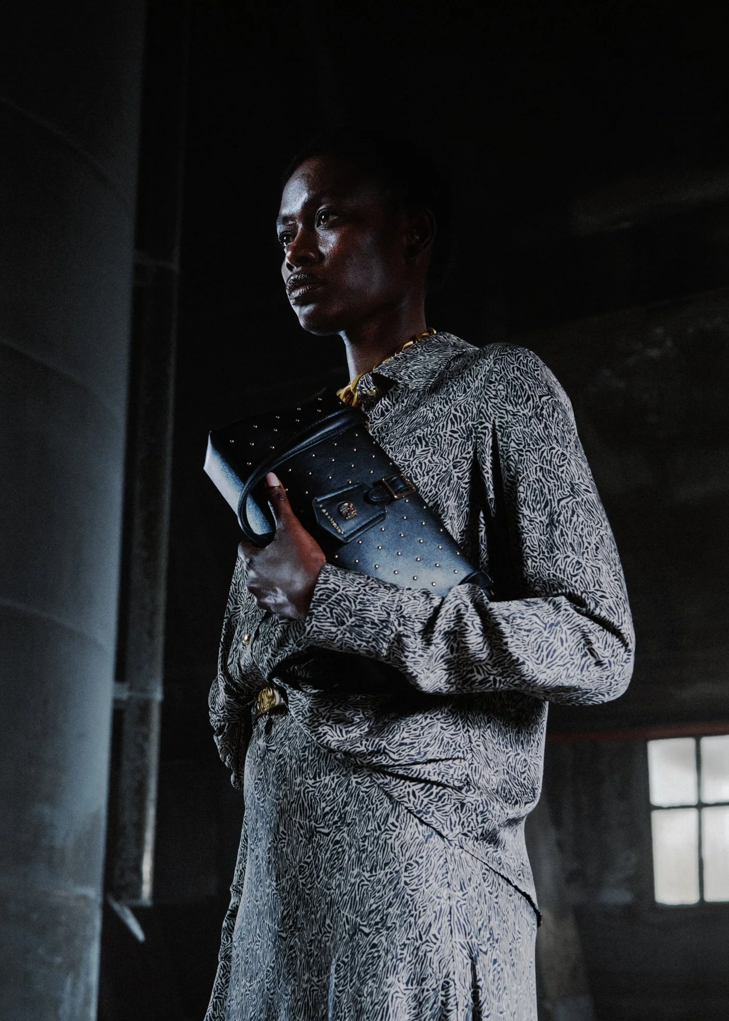 Una donna vestita con un completo a fantasia bianca e nera, che tiene un clutch nero con borchie, in un ambiente scuro.