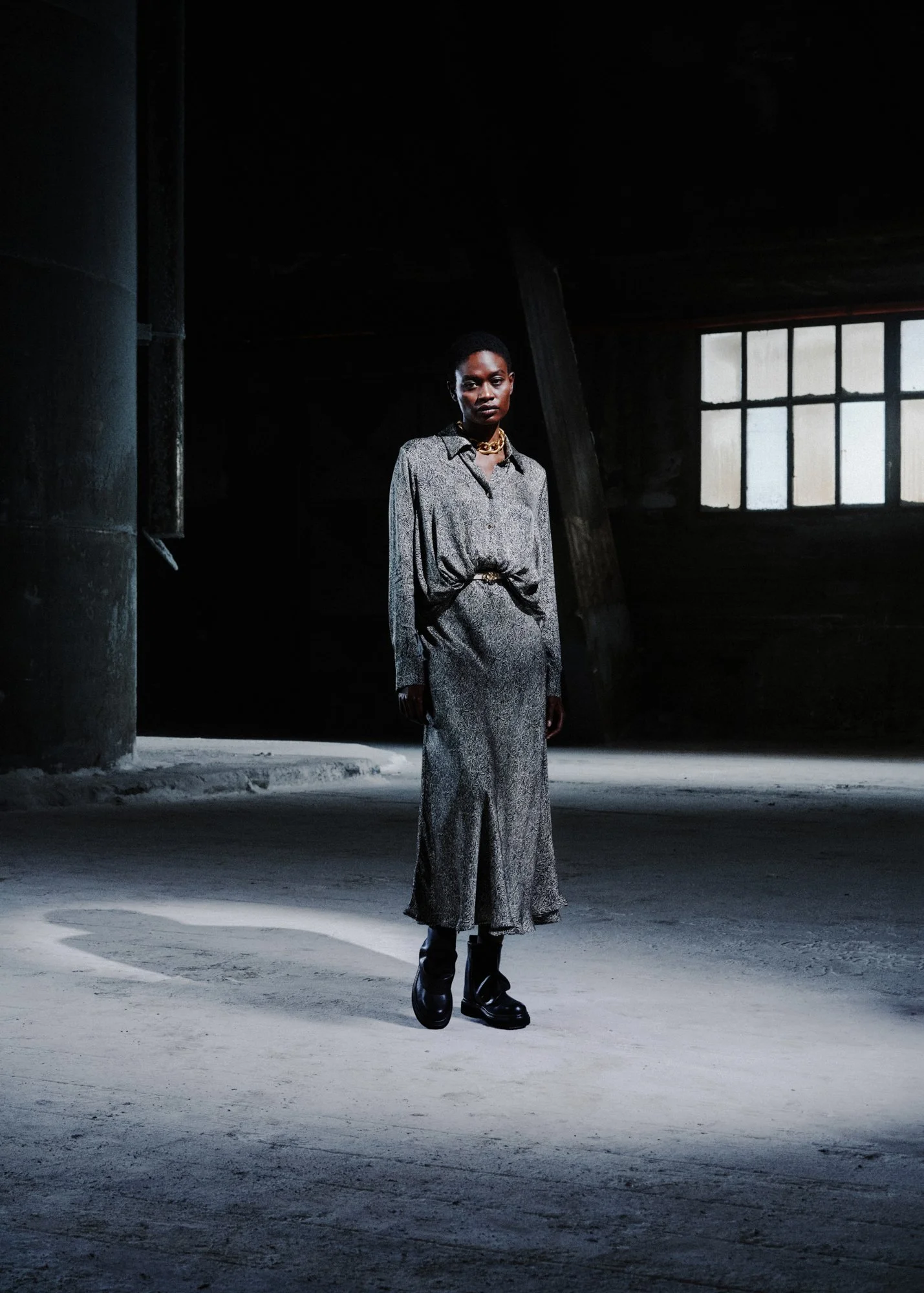Una donna vestita con un completo a fantasia grigia, con un cappotto lungo e tacchi, si trova in un ambiente industriale abbandonato con pareti di mattoni e una grande finestra con illuminazione naturale.