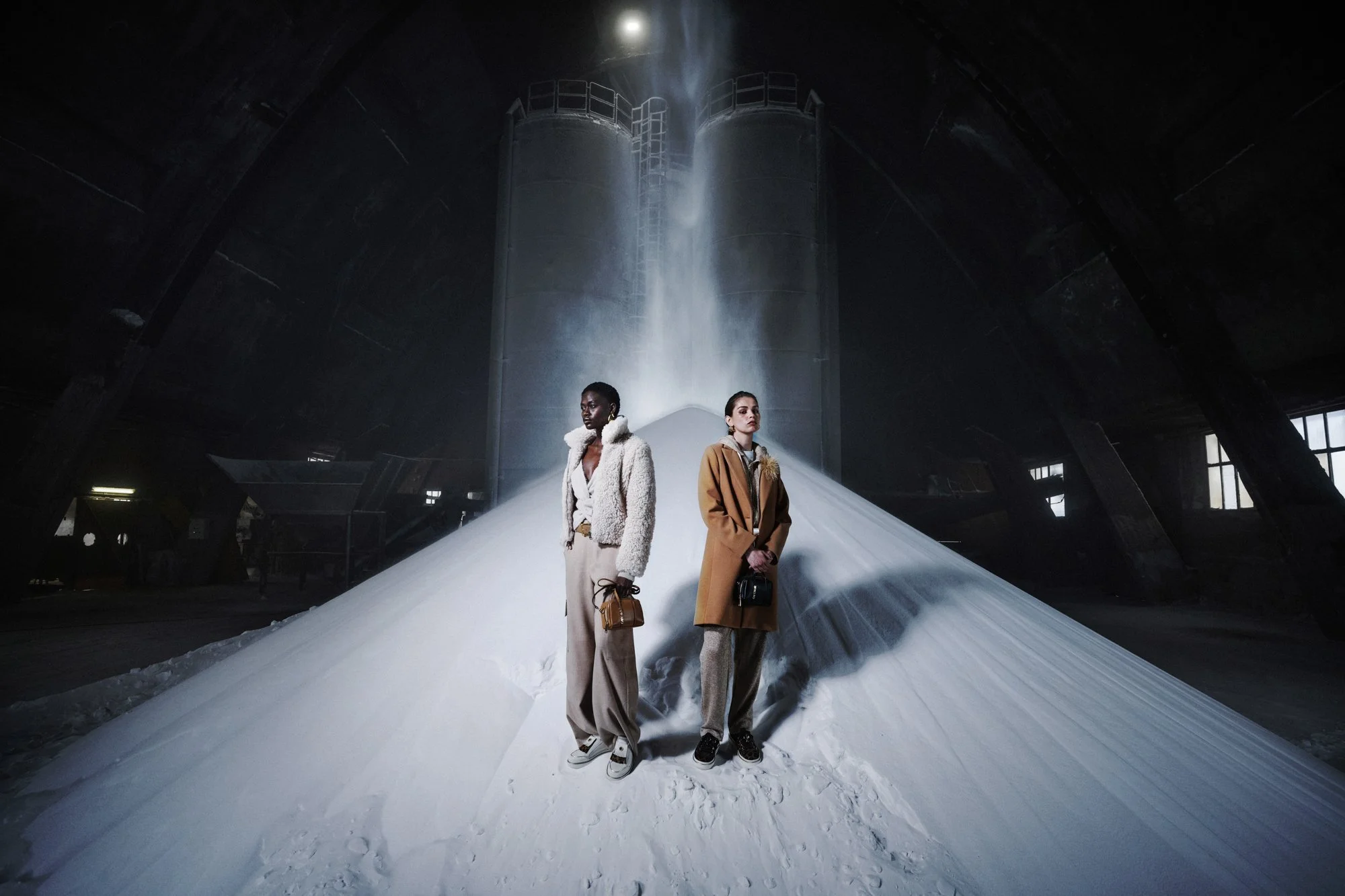 Due donne in abbigliamento invernale si trovano in un ambiente industriale, davanti a una pila di neve o materiale bianco, con grandi strutture di metallo sullo sfondo e luce che filtra dall'alto.