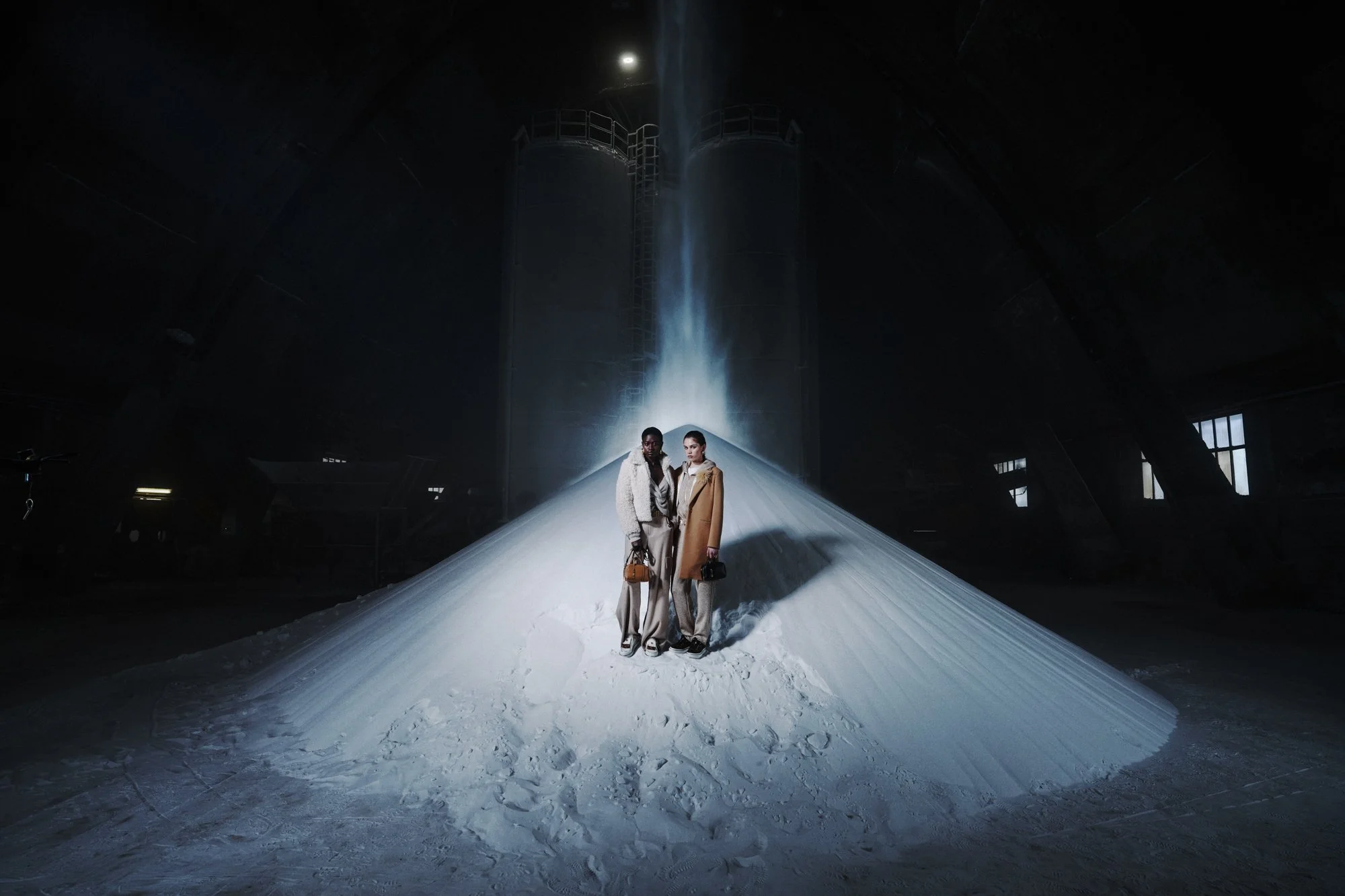 Due donne in abiti eleganti e scarpe da ginnastica posano su una grande montagna di neve all'interno di un edificio industriale con tubature e strutture di acciaio visibili.