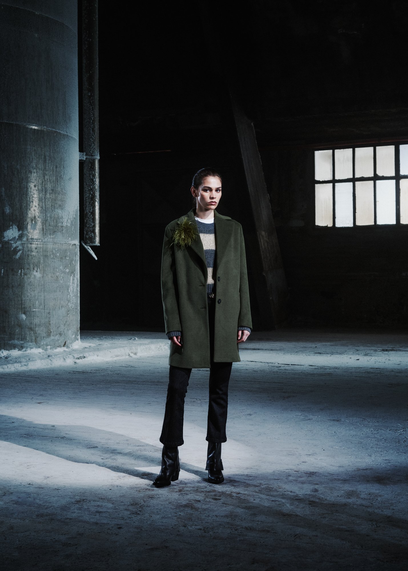 Una donna indossa un cappotto verde con una spilla a forma di muschio e pantaloni neri in un ambiente industriale oscuro con grandi finestre e muri di cemento.