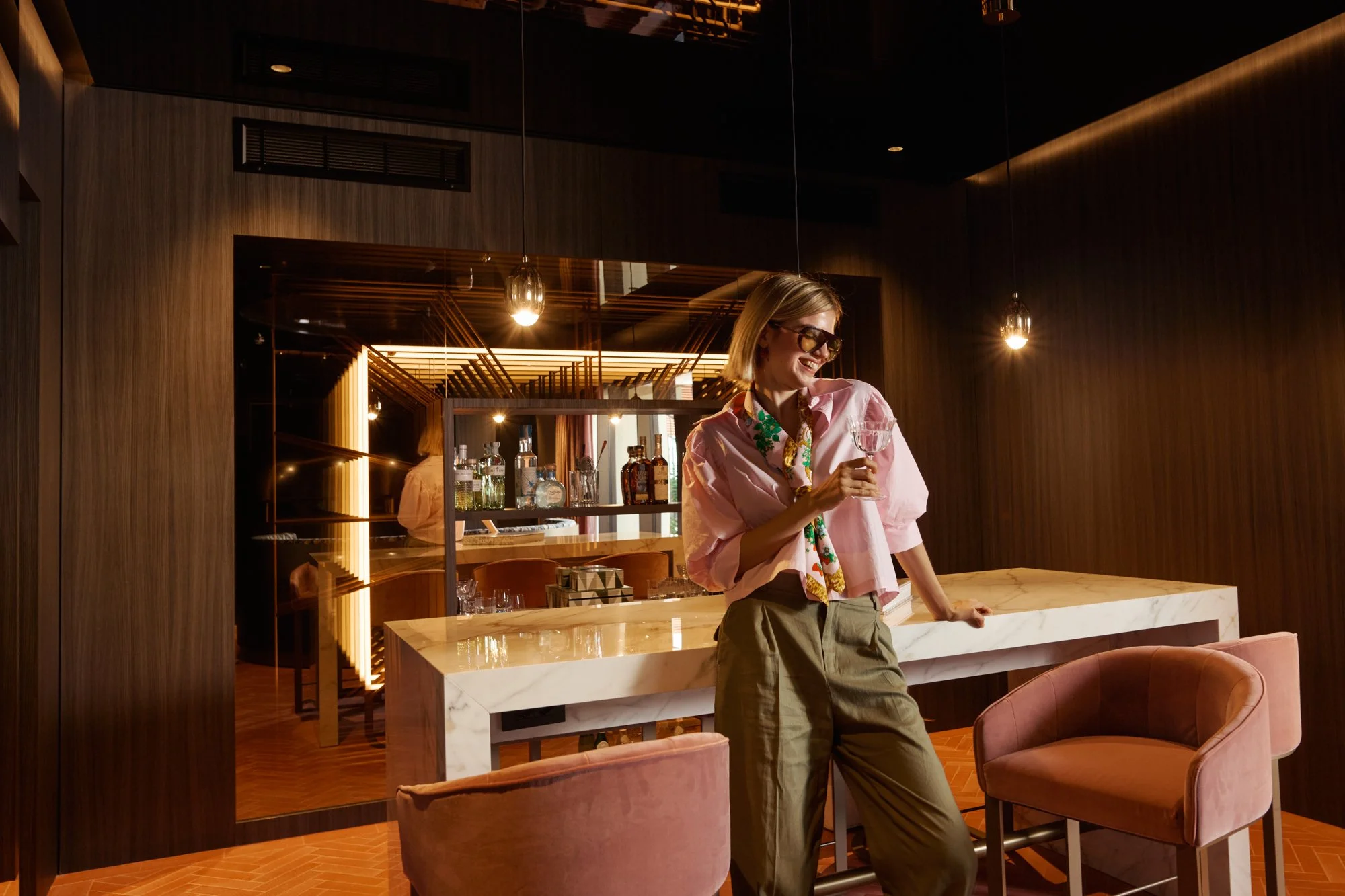 Donna sorridente con occhiali da sole, cropped, indossa una camicia rosa e pantaloni verdi, mentre sorseggia un bicchiere di cocktail nel bar di un ristorante elegante con arredi in legno e lampade luminose