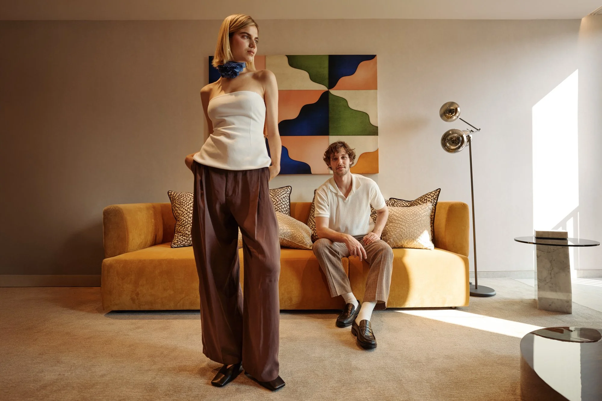 Una donna seduta su un divano arancione, un uomo con pantaloni beige e camicia bianca seduto di fronte, un quadro astratto alle spalle, una lampada moderna vicino alla finestra che entra luce naturale.