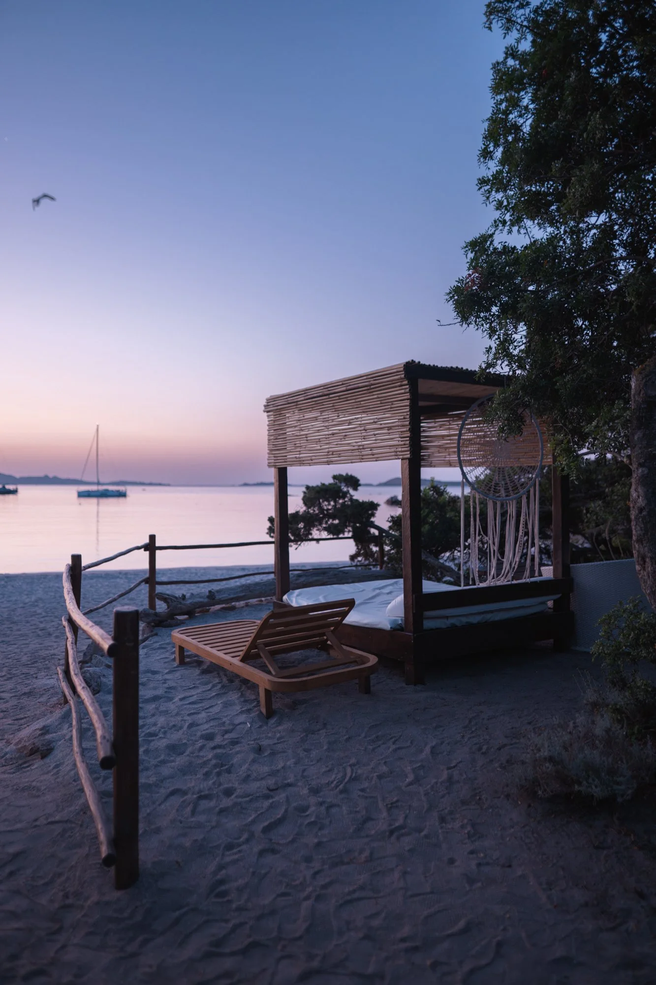 Zona relax sulla spiaggia con lettino e cabina di legno, vista sul mare al tramonto con barche all'orizzonte, albero e gabbiano nel cielo.