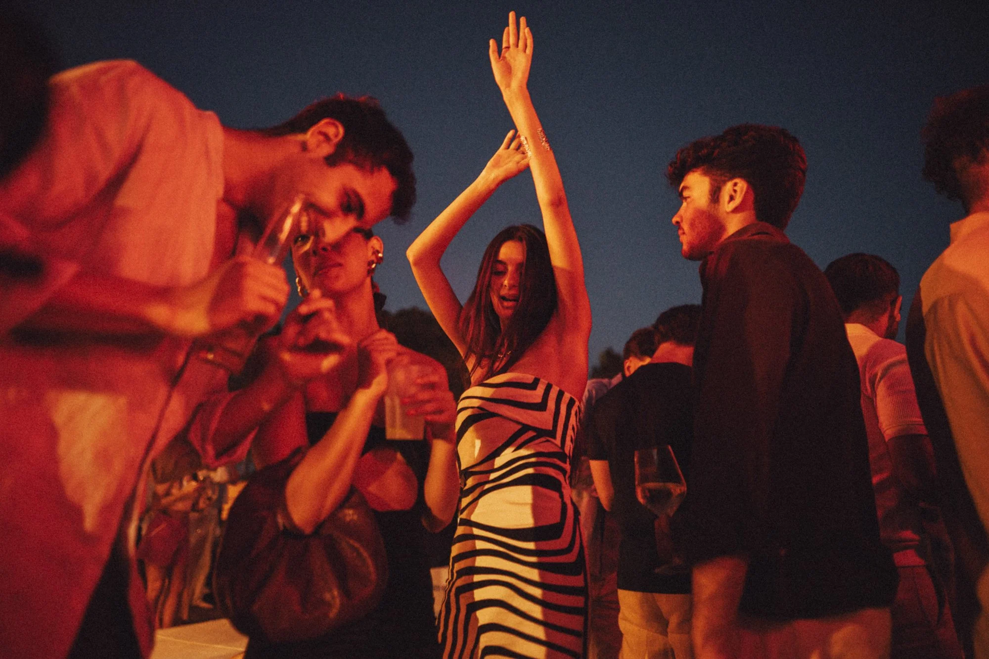 Gruppo di persone in un evento sociale, alcune che ballano e altre che parlano, sotto un cielo al tramonto o di sera, con luci calde e atmosfera vivace.