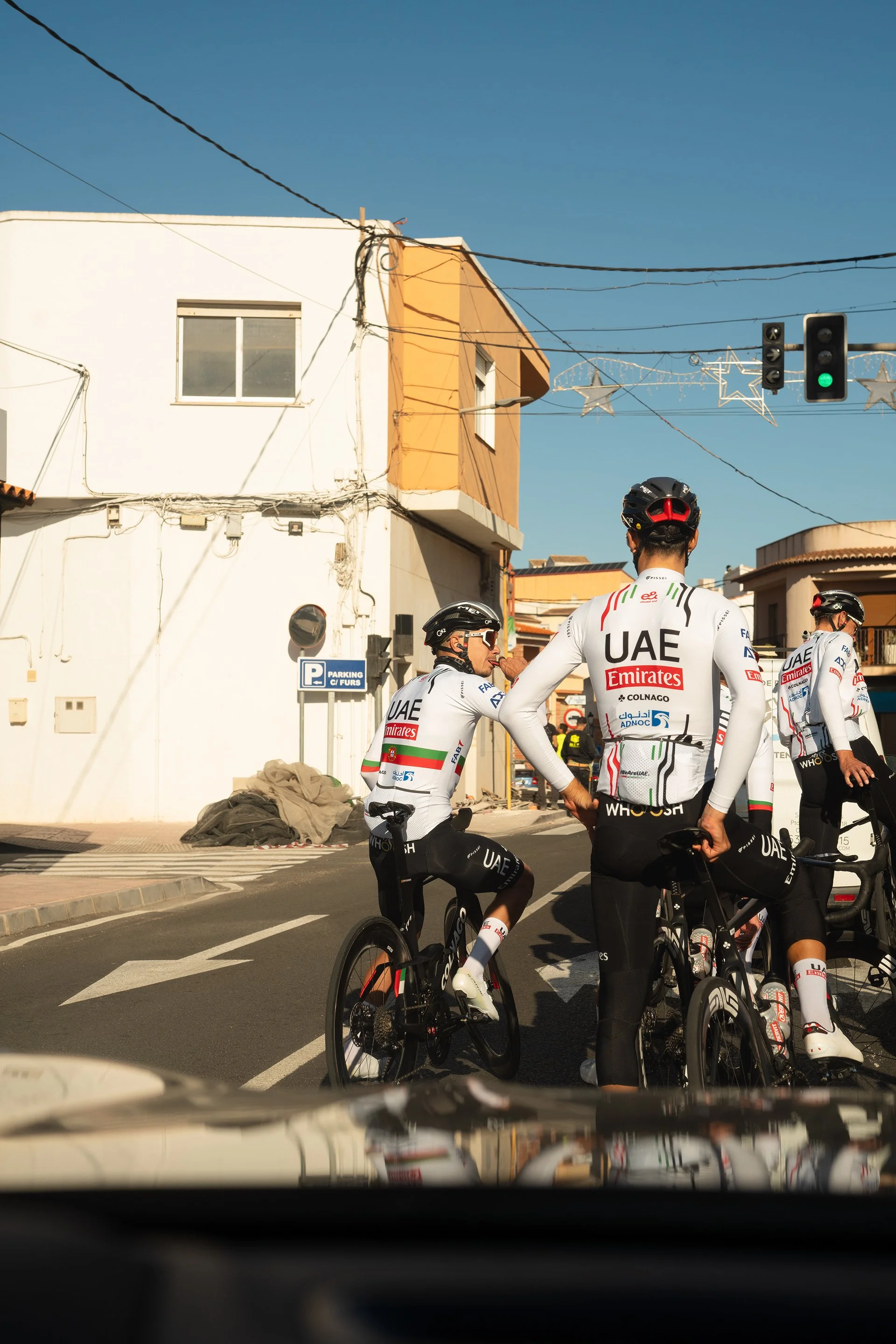 Gruppo di ciclisti vestiti con divise della squadra UAE Emirates, alcuni impegnati in conversazione, si trovano in un'area urbana con edifici colorati, decorazioni natalizie e un semaforo verde.
