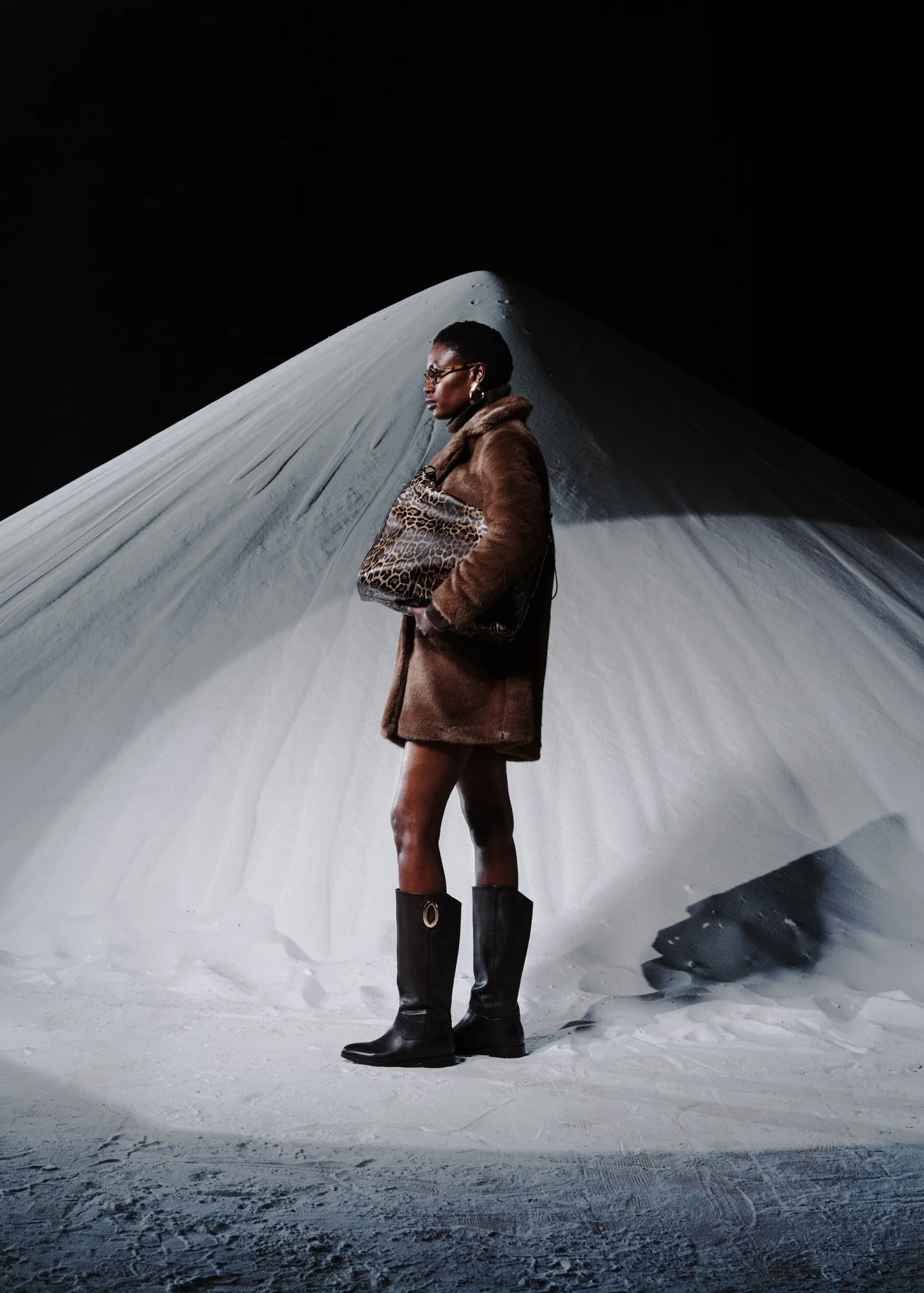 Donna con cappotto marrone, pantaloni corti e stivali neri, porta una borsetta animalier, si trova su una superficie di neve con una montagna dietro e un cielo nero.