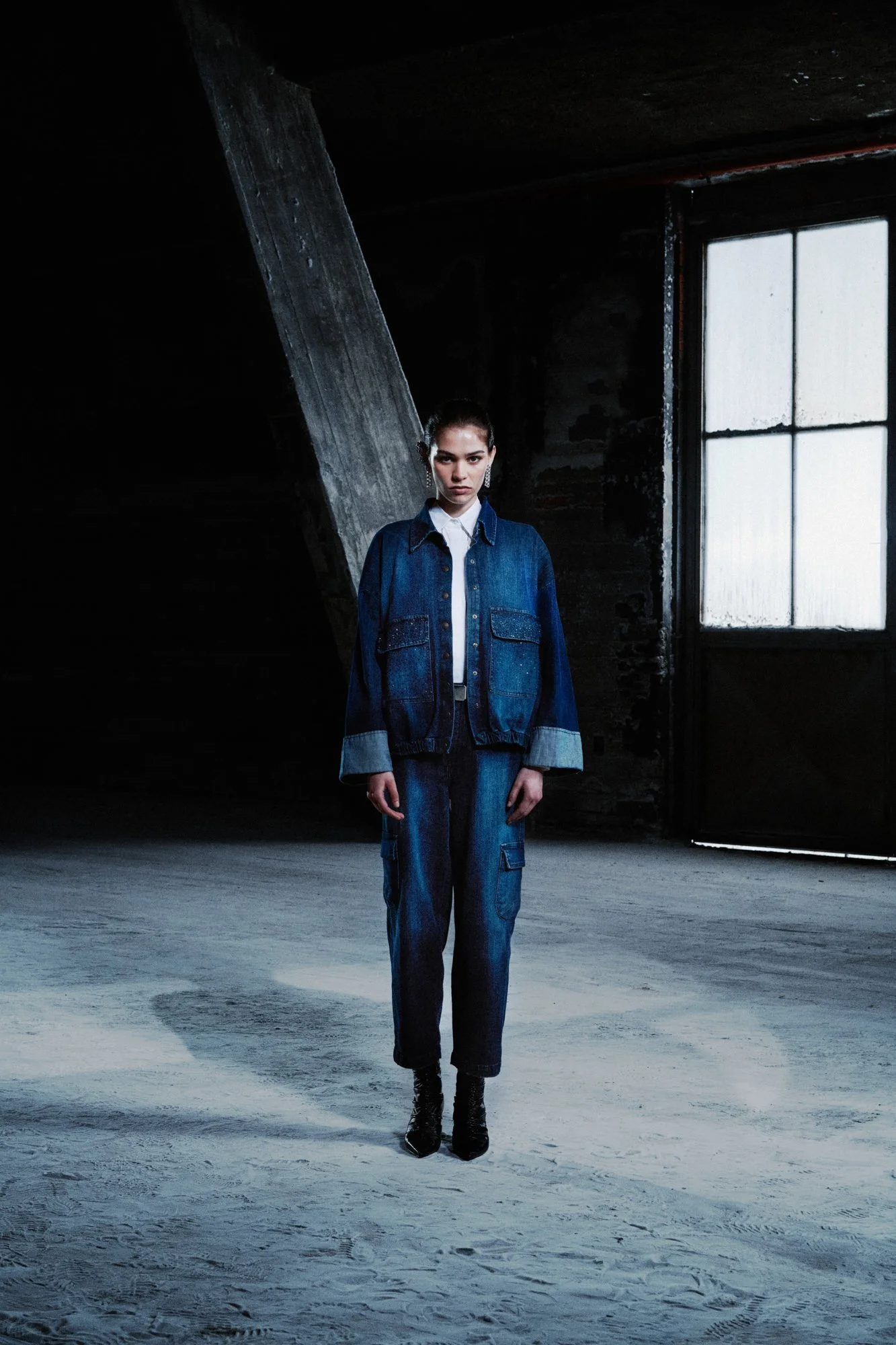 Una donna vestita con un completo di jeans scuri, stivali neri, occhiali e orecchini, si trova in un ambiente industriale oscuro con pareti di mattoni e un'ampia finestra lucida.